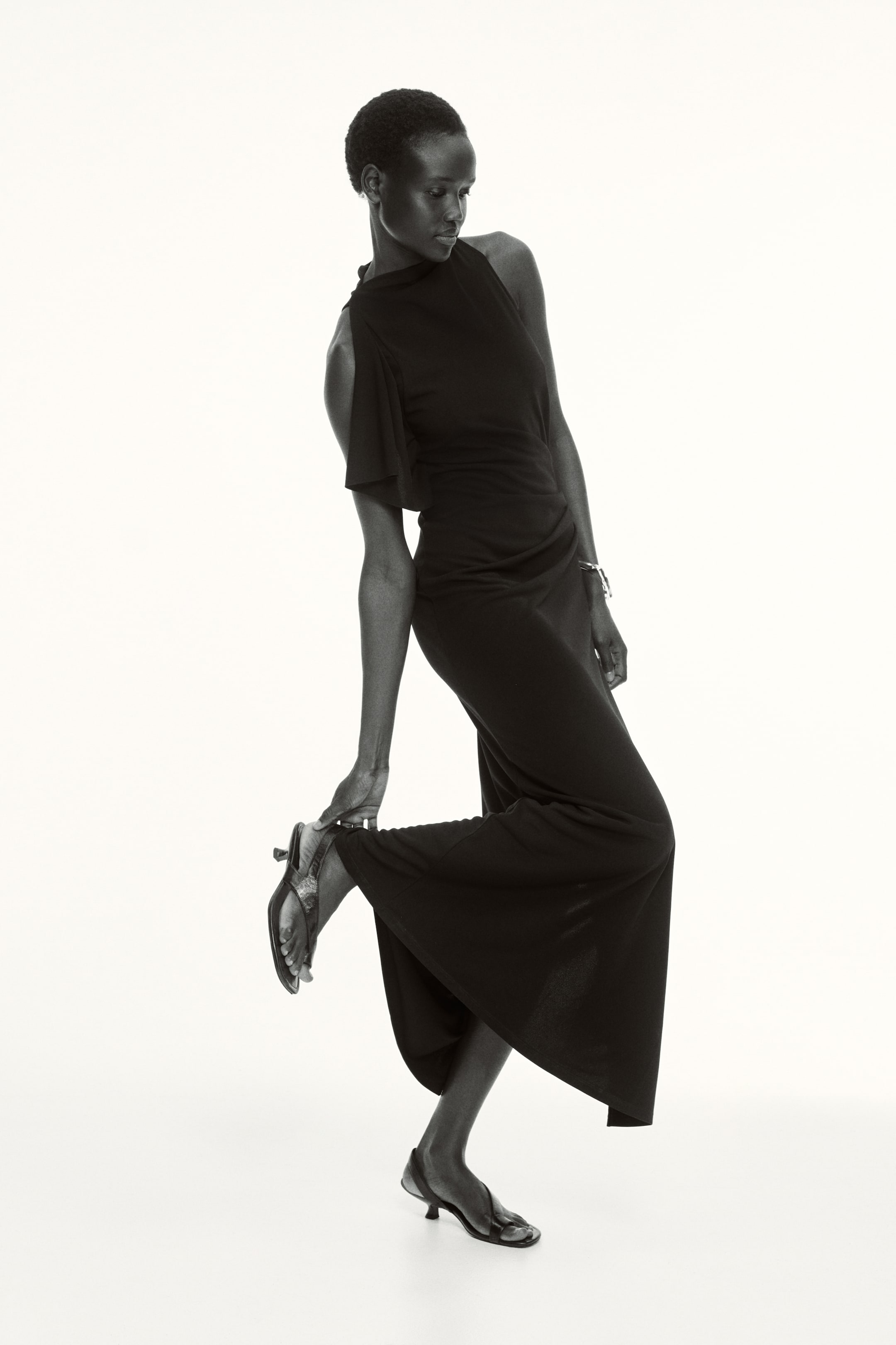 Visa större bild: Den svarta jumpsuiten med vida ben har en asymmetrisk halsringning med en bar axel och den andra prydd med en bred, draperad ärm. Den övergår i vida, avslappnade byxor och skapar en flytande silhuett.