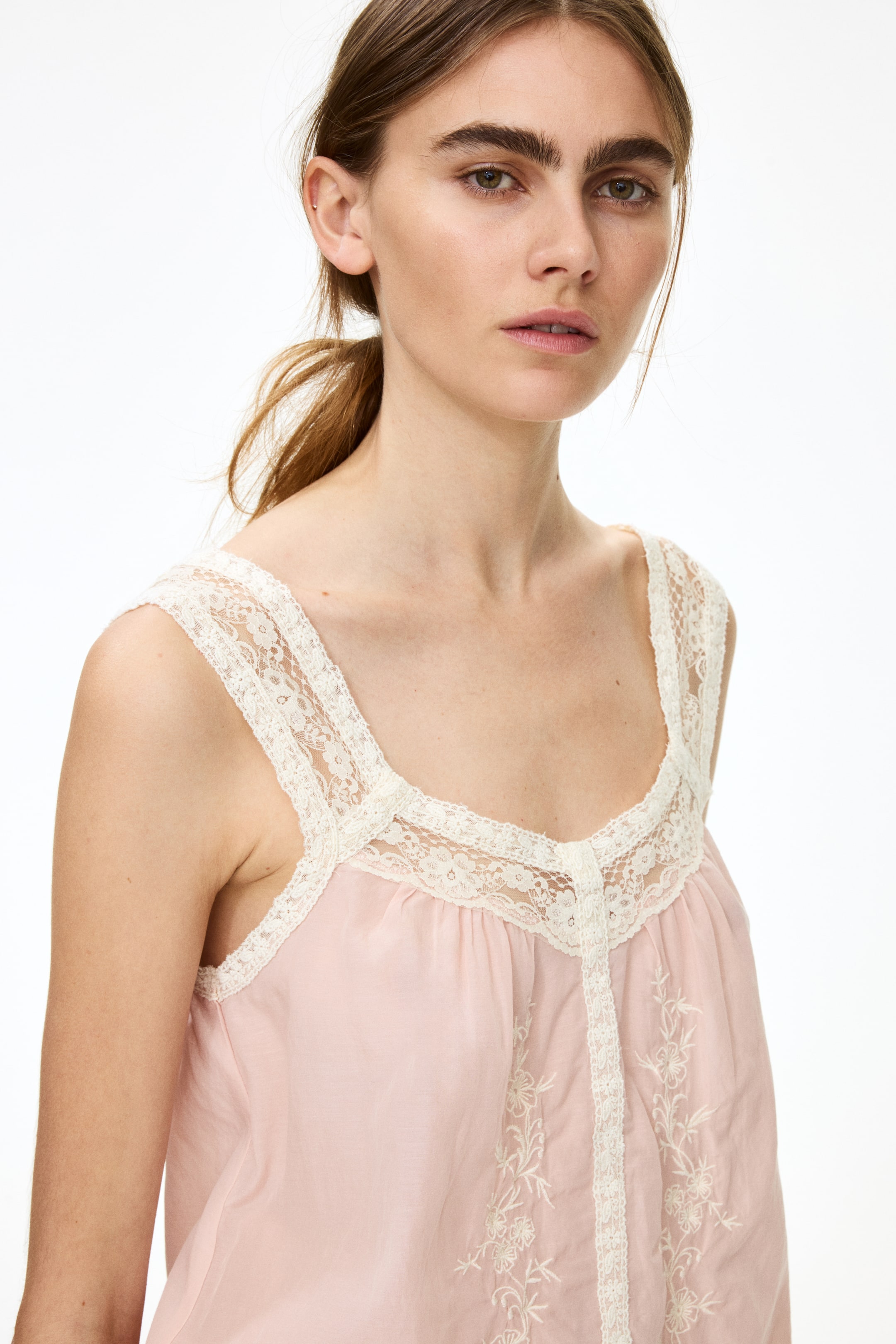 View larger image: A light pink camisole featuring off-white lace trim on its wide straps, square neckline, and down the center front, where the fabric is gathered and white floral embroidery details the lower panel.