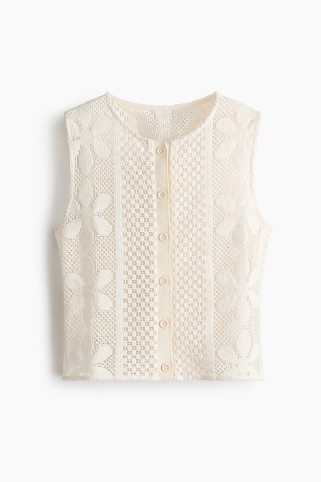 Eine cremefarbene, ärmellose Strickjacke zeigt ihre Vorderseite und verfügt über eine filigrane Spitzenstruktur mit abwechselnden floralen und geometrischen Mustern. Sie ist mit einem runden Halsausschnitt gearbeitet und hat eine zentrale Knopfleiste, die mit sieben sichtbaren, runden Knöpfen geschlossen wird. - 1