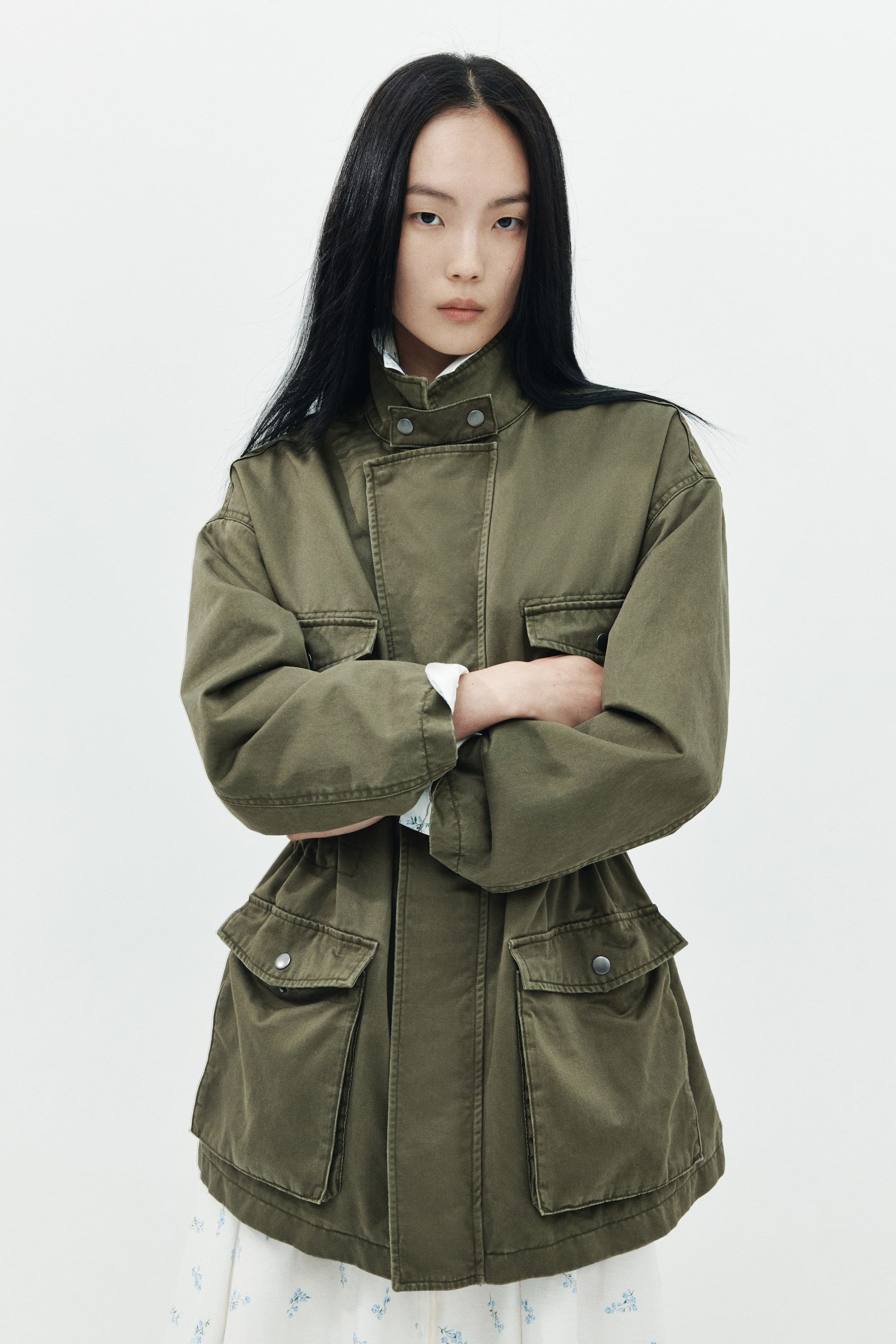 이미지 확대 보기: 모델이 팔짱을 낀 포즈를 취하며 올리브 그린 컬러의 유틸리티 재킷을 착용하고 있습니다. 재킷은 스냅 여밈이 있는 하이 스탠드 칼라, 히든 프런트 플래킷, 네 개의 큰 플랩 포켓이 특징이며, 칼라, 커프스, 밑단에서 보이는 화이트 패턴 의상 위에 레이어드되어 있습니다.