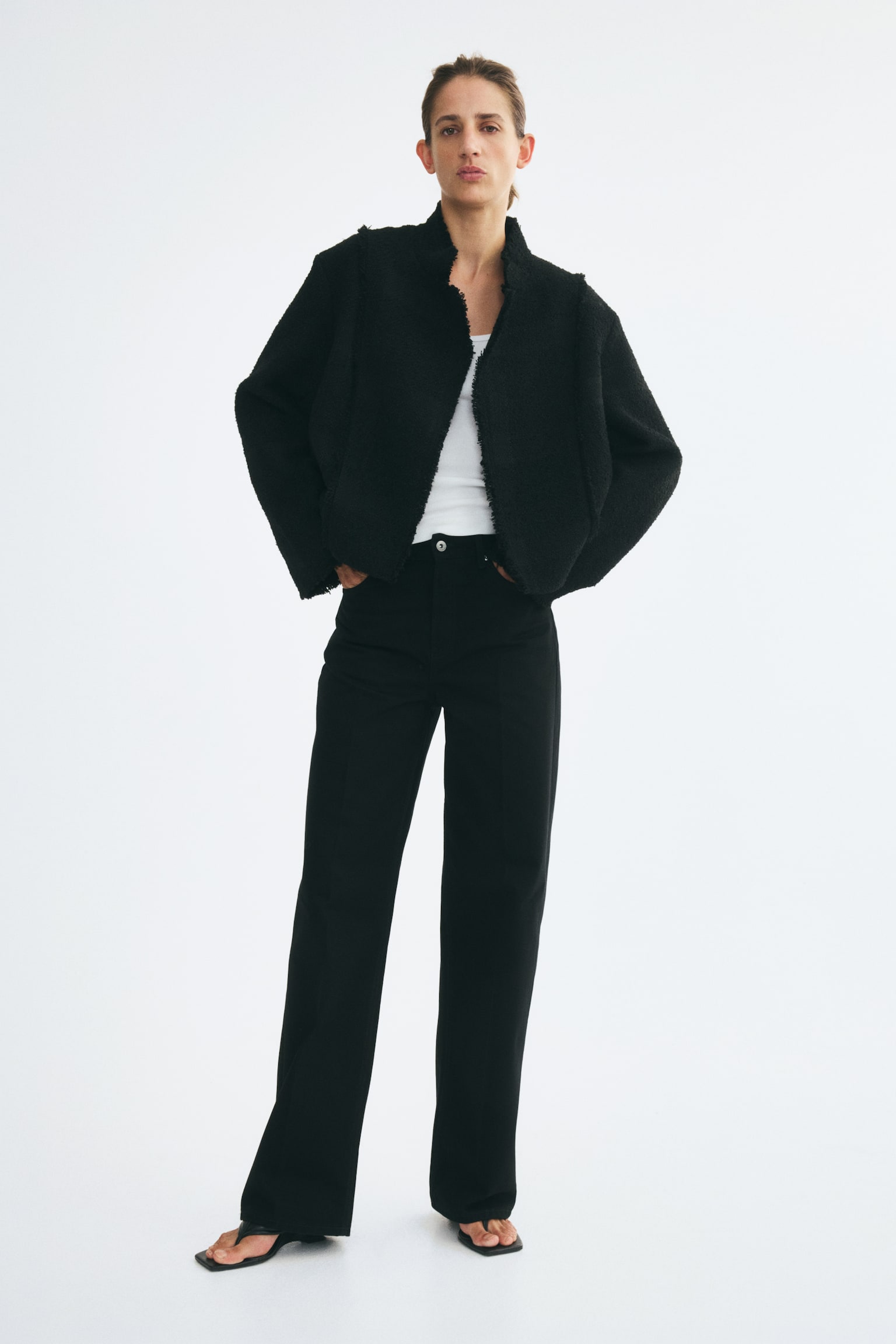 A black cropped jacket features a textured, boucle-like fabric and a front-facing design with a high stand collar. It has an asymmetrical front overlap, wide long sleeves, and raw, frayed edges along the seams, collar, cuffs, and hem. - 1
