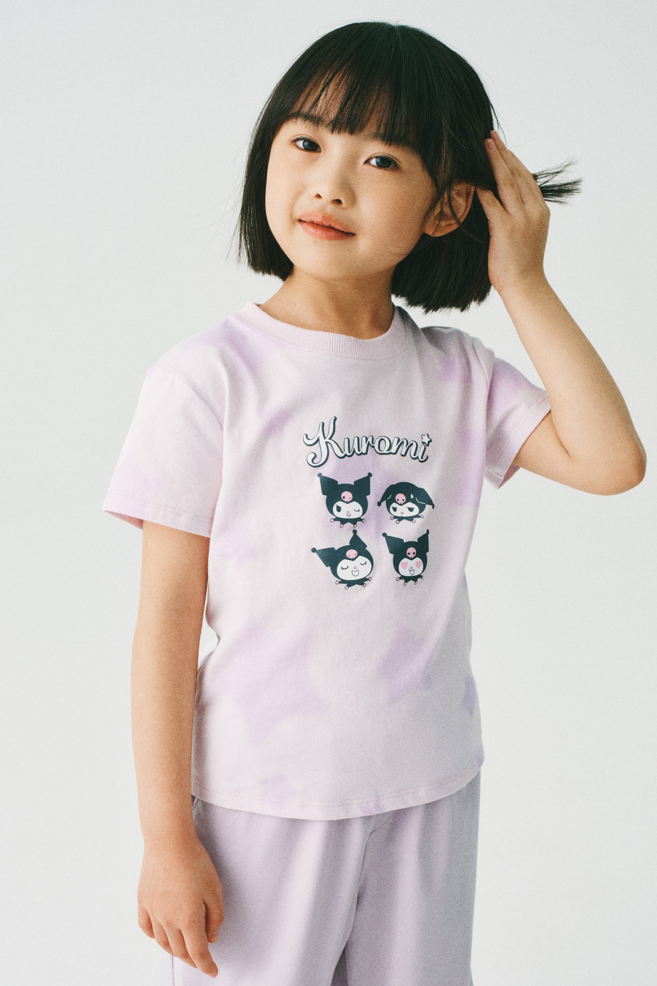 Xem ảnh lớn hơn: Người mẫu đang mặc một chiếc áo thun cotton jersey màu tím nhạt in hình, có cổ tròn bo gân và tay ngắn. Áo thun có thiết kế chữ "Kuromi" và bốn hình ảnh nhân vật hoạt hình ở phía trước.