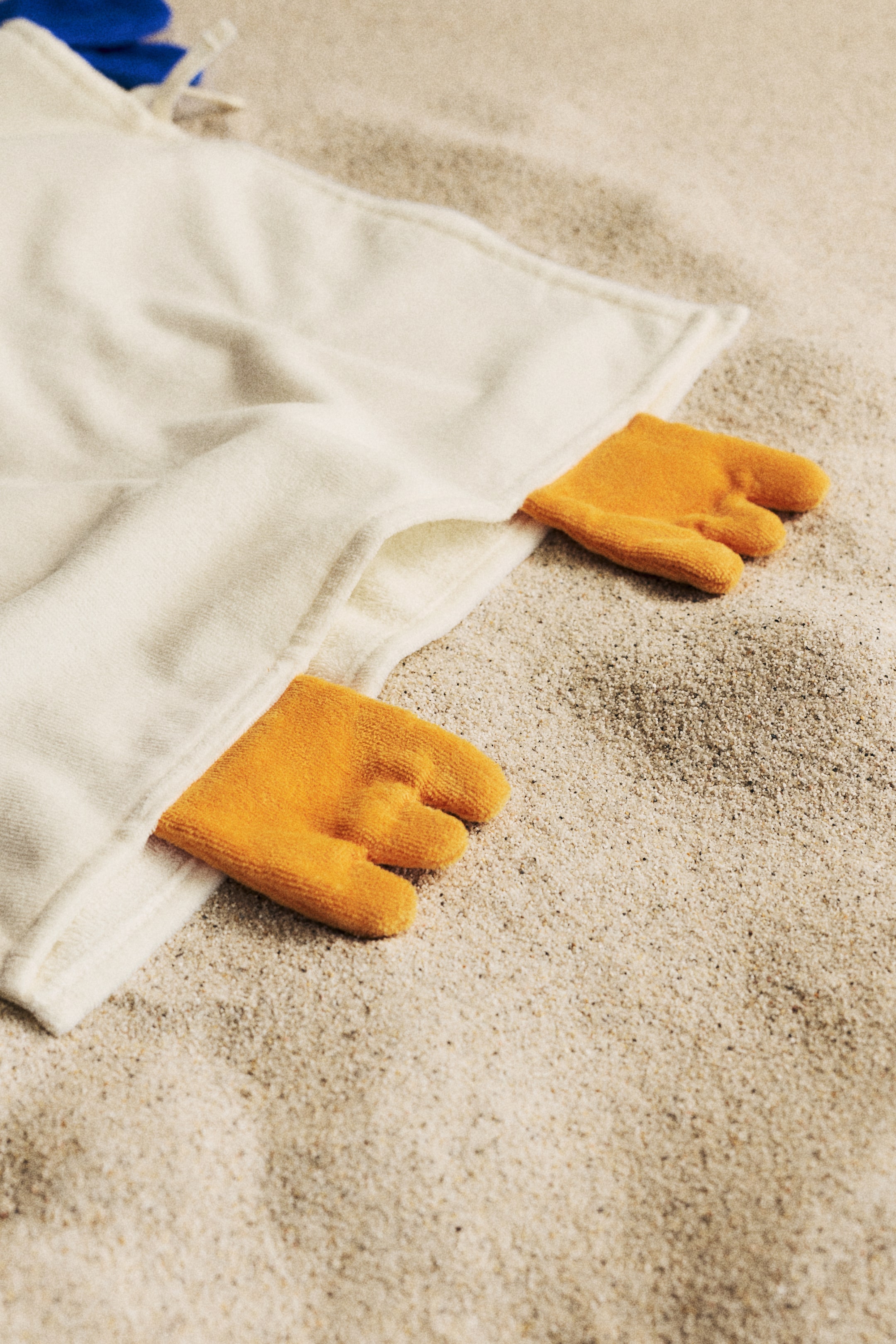 Zobacz większy obraz: Ręcznik z tkaniny frotte w kolorze kremowym, ozdobiony dwoma pomarańczowymi pięciopalczastymi wyrostkami. Wyrostki wyłaniają się z fałd materiału ręcznika i są skierowane w dół.