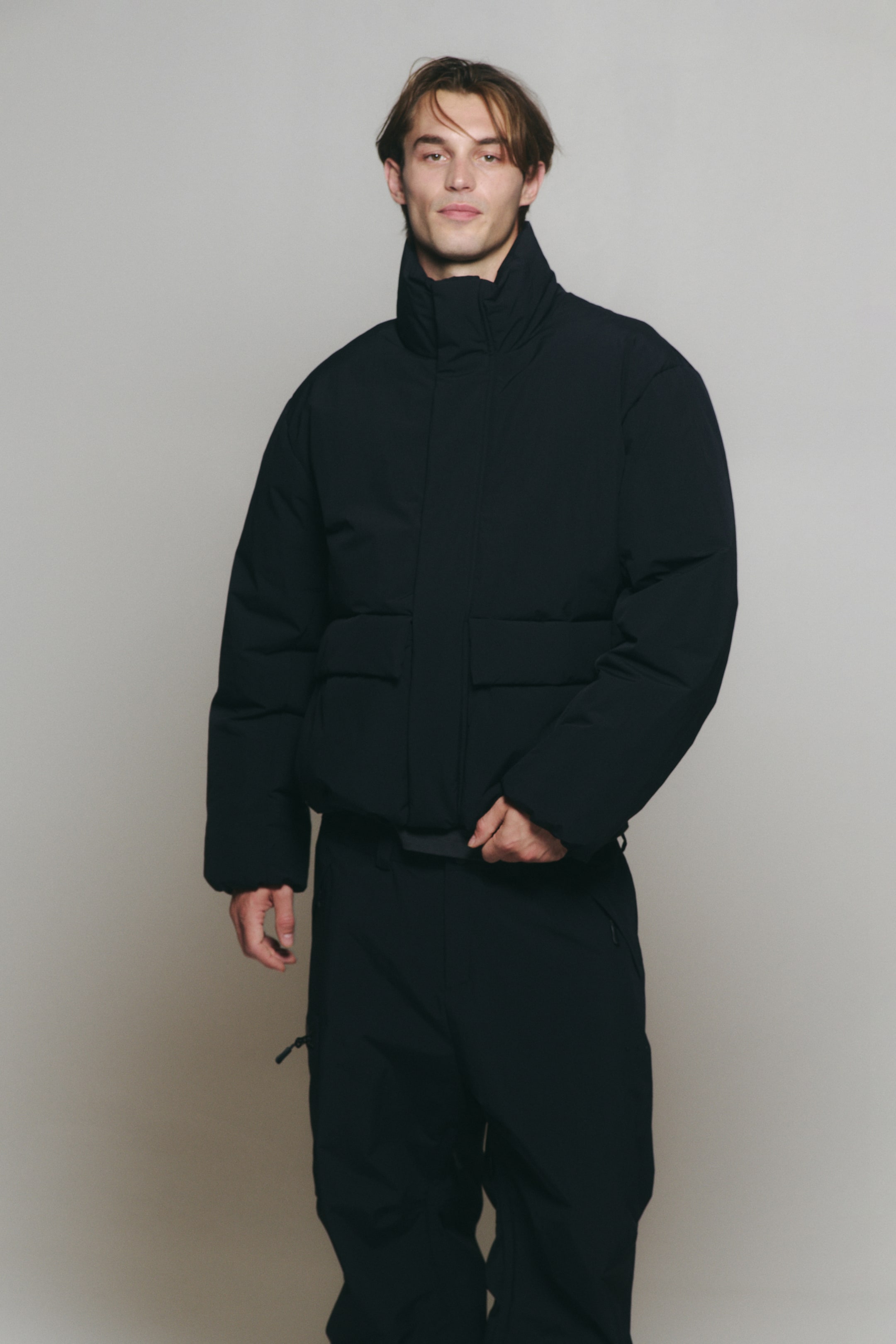View larger image: A black, high-collar puffer jacket with large front flap pockets is being worn by the model, paired with matching black utility pants.