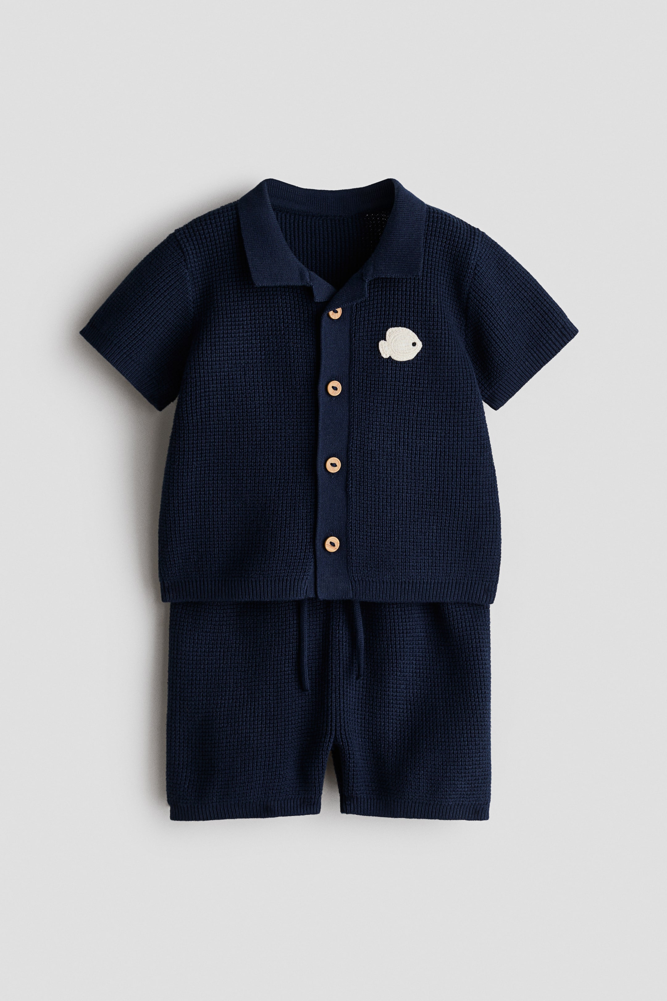 View larger image: A 2-piece navy blue cotton waffle-knit set is showcasing a short-sleeved top and matching shorts, viewed from the front. The top features a collar, a five-button placket with light-colored buttons, and a white embroidered fish appliqué on the left chest, while the shorts exhibit a visible drawstring waist.
