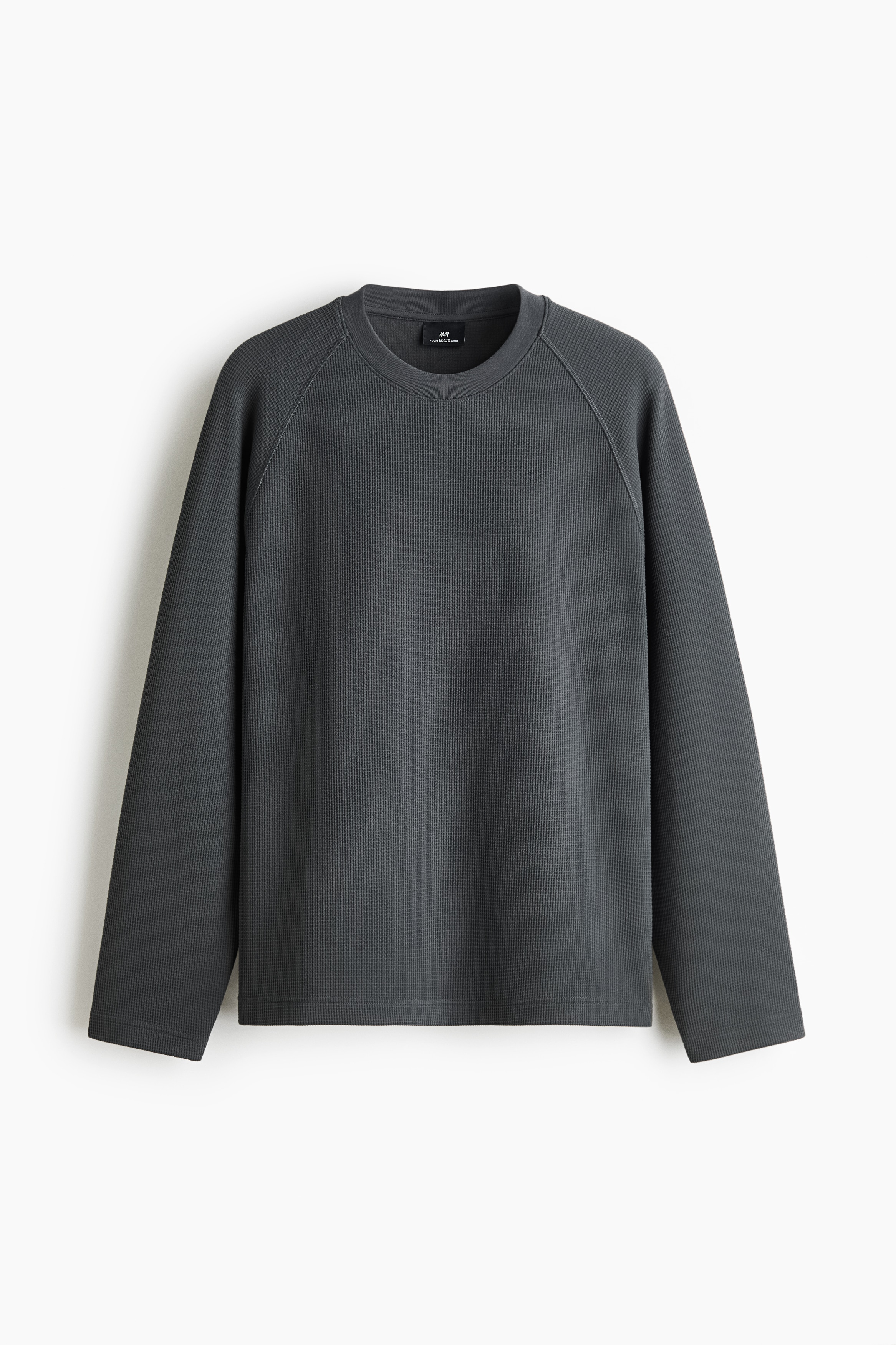 Le mannequin porte un tee Ã  manches longues gris foncÃ© en jersey gaufrÃ©, Ã  coupe dÃ©contractÃ©e, avec un col rond et des manches raglan. Le mannequin est debout, les mains dans les poches dâun pantalon gris pÃ¢le, mettant en valeur lâourlet droit du haut.