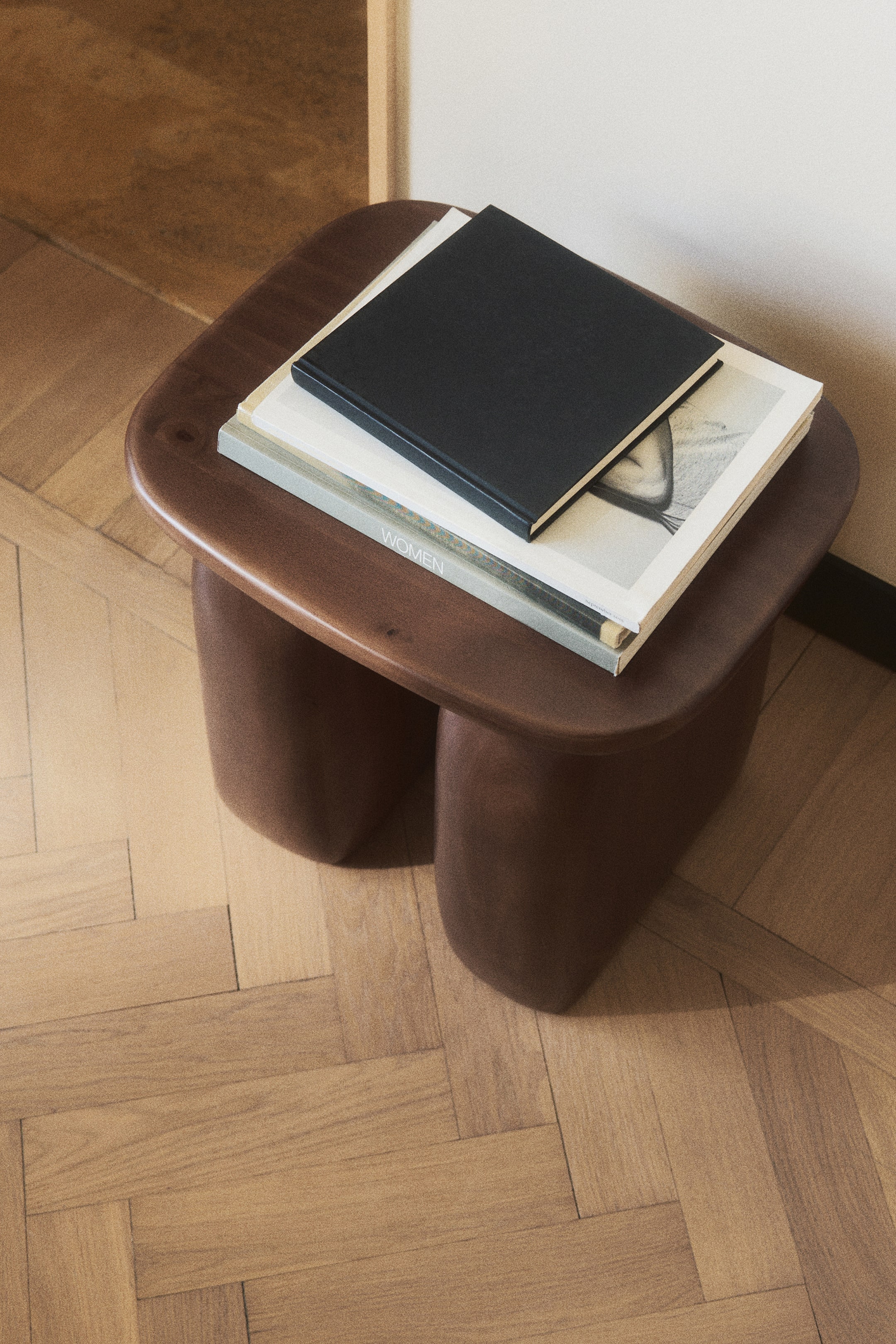 Grotere afbeelding bekijken: Een donkerbruine houten bijzettafel met een glad, afgerond rechthoekig blad, gedragen door dikke, organisch gevormde poten. Op het tafelblad ligt een stapel van drie boeken.