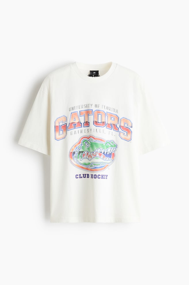 En kremfarget T-skjorte med rund hals er avbildet forfra, med avslappet passform og korte ermer. Forsiden viser en slitt collegiate-grafikk med teksten «UNIVERSITY OF FLORIDA GATORS» buet over en grønn alligatorlogo og «CLUB HOCKEY» trykt under. - 1