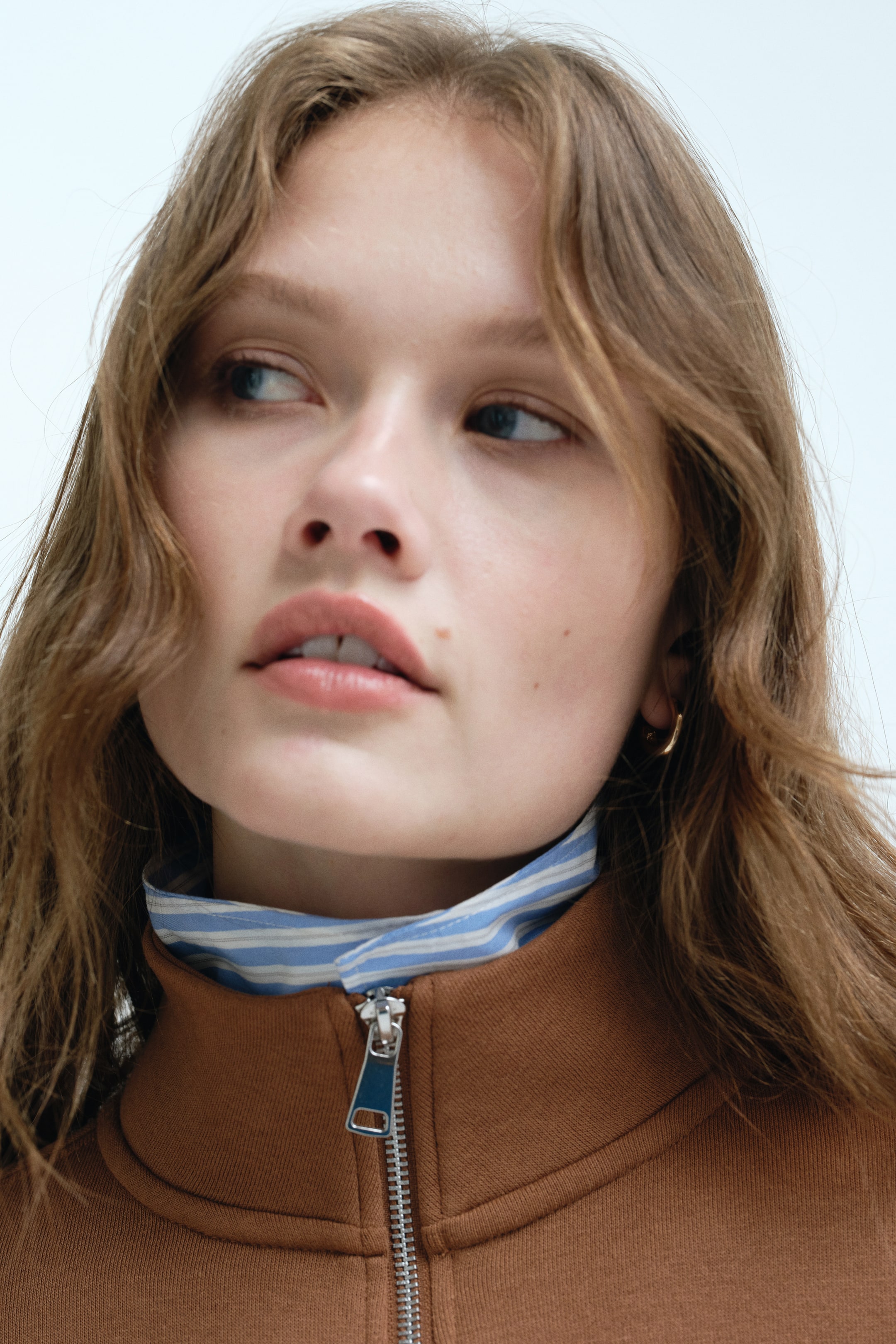 View larger image: A brown high-neck jacket features a partially unzipped silver zipper, revealing a blue-and-white striped shirt collar underneath.
