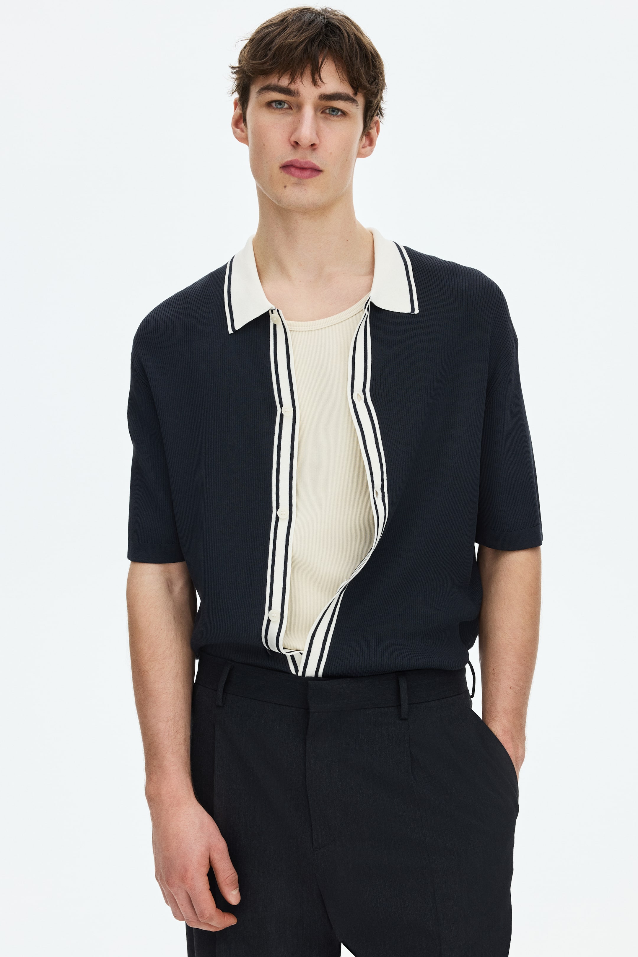 View larger image: A navy blue ribbed knit short-sleeve polo shirt is shown facing forward, featuring an off-white collar accented with a navy stripe. The front placket, adorned with off-white and navy vertical stripes, is unbuttoned, layered over a light beige ribbed top.