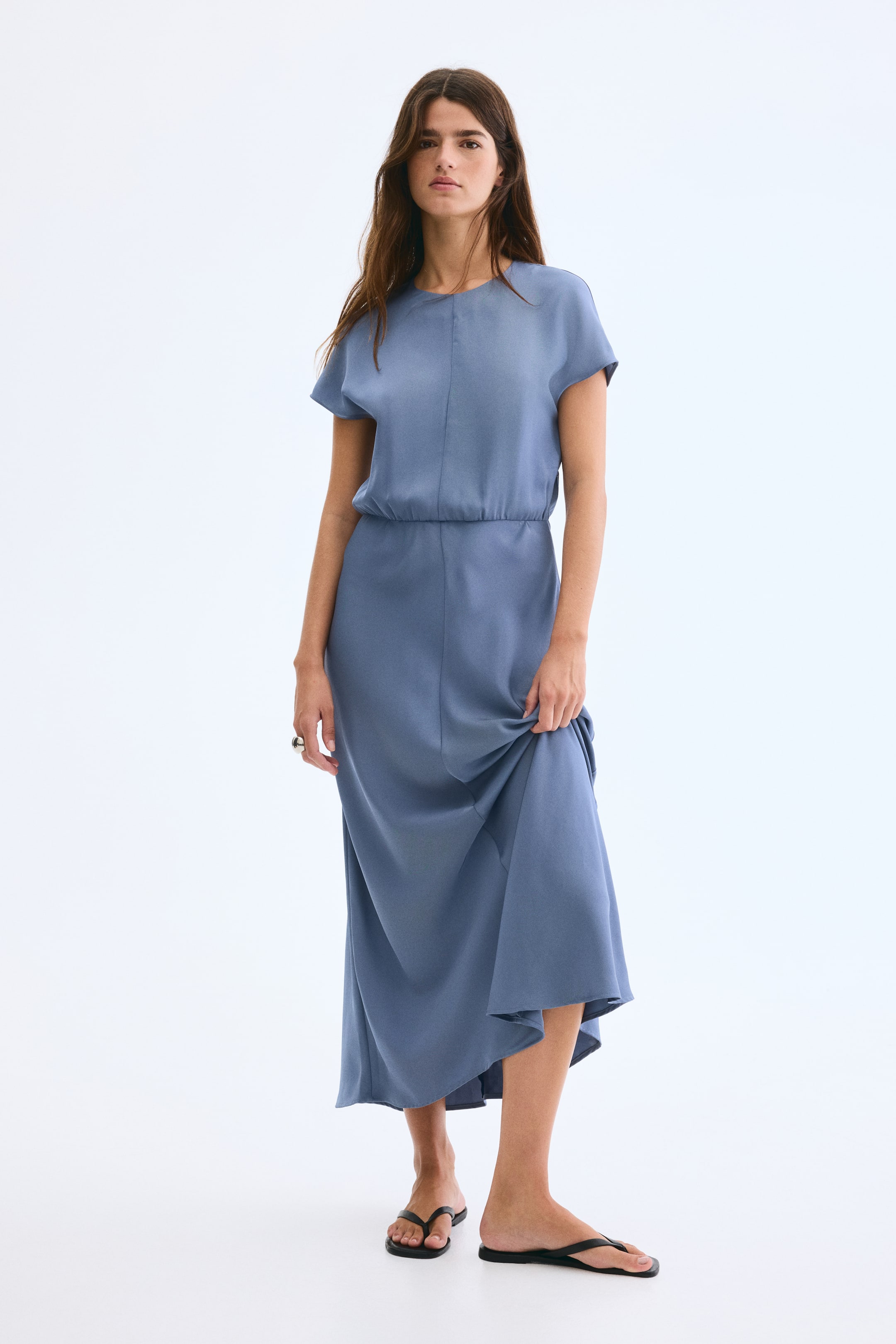 View larger image: A blue midi dress is being presented front-facing, featuring a round neckline and cap sleeves. The dress is cinched at the waist with elastication, and its flowing skirt is extending to mid-calf, with a hand gently lifting a portion of the hem.