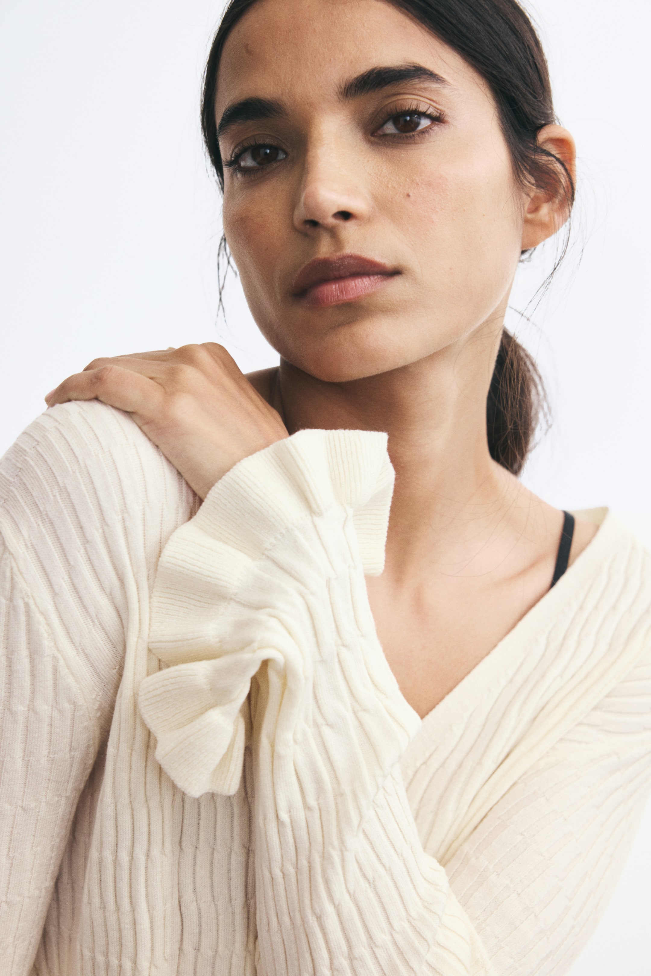 Ver imagem maior: Um cardigan em malha texturada, branco sujo, com decote em V, está a ser usado pela modelo. A modelo está a pousar uma mão no ombro, destacando o detalhe do punho com folho da manga comprida.