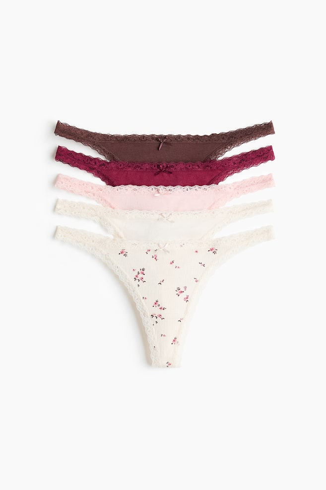Ein Fünferpack niedrig geschnittener G-Strings aus Baumwolljersey ist in einer vertikalen Stapelanordnung von vorne zu sehen und zeigt die verschiedenen Designs. Die Auswahl umfasst die unifarbenen Varianten in Dunkelbraun, Burgunderrot, Hellrosa und Creme sowie ein geripptes Paar in Off-White mit rosa Blumenmuster, jeweils mit zarten Spitzenabschlüssen und einer kleinen Schleife in der Mitte. - 1
