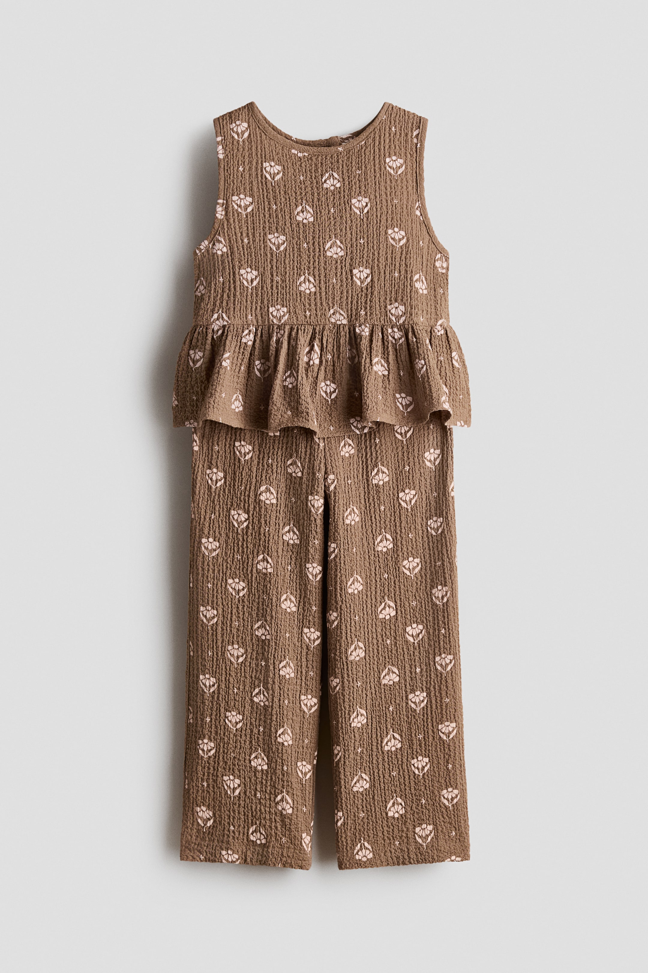 View larger image: A front view of a two-piece outfit features a brown crinkled texture with a repeating light pink stylized floral pattern. The sleeveless top has a round neckline and a gathered peplum hem, while the matching pants showcase a straight-leg cut.