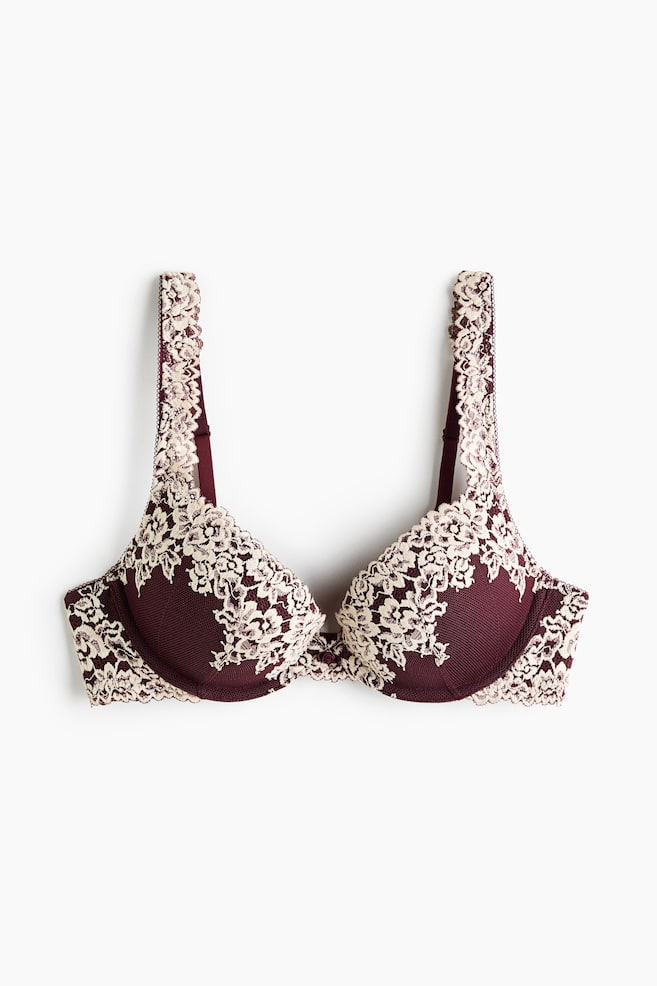 Un soutien-gorge super push-up bordeaux et crÃ¨me est prÃ©sentÃ© de face, mettant en valeur des bonnets rembourrÃ©s Ã  armatures avec une fine texture en mesh. Le modÃ¨le se distingue par de superpositions de dentelle crÃ¨me travaillÃ©es qui soulignent les bonnets, la basque et les larges bretelles. - 1