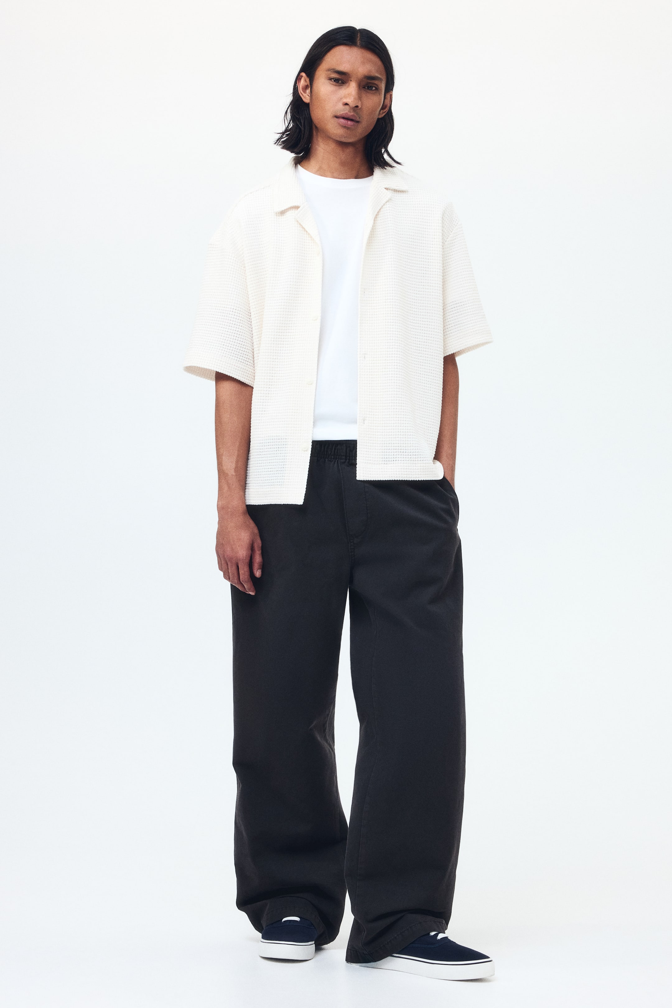 View larger image: The ensemble features a cream short-sleeve shirt with a waffle-knit texture and camp collar, worn open over a plain white crew-neck t-shirt. Dark gray wide-leg trousers with an elastic waistband are paired with navy canvas sneakers sporting white soles. The outfit is shown facing forward, highlighting its relaxed fit and design.