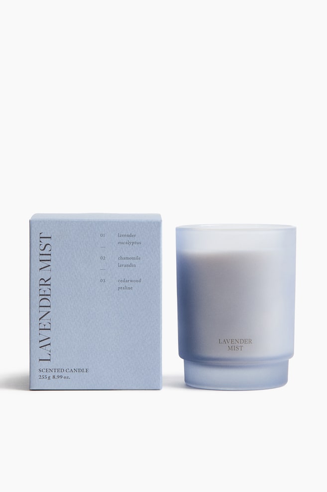 ラベンダーカラーの長方形の箱が立ち、正面を向いています。箱の前面には「LAVENDER MIST」と香りのノートが縦に表示されています。その隣には、白いワックスが入ったフロスト加工のラベンダー色の円筒形ガラスキャンドルジャーが立ち、正面に「LAVENDER MIST」と表示されています。 - 1