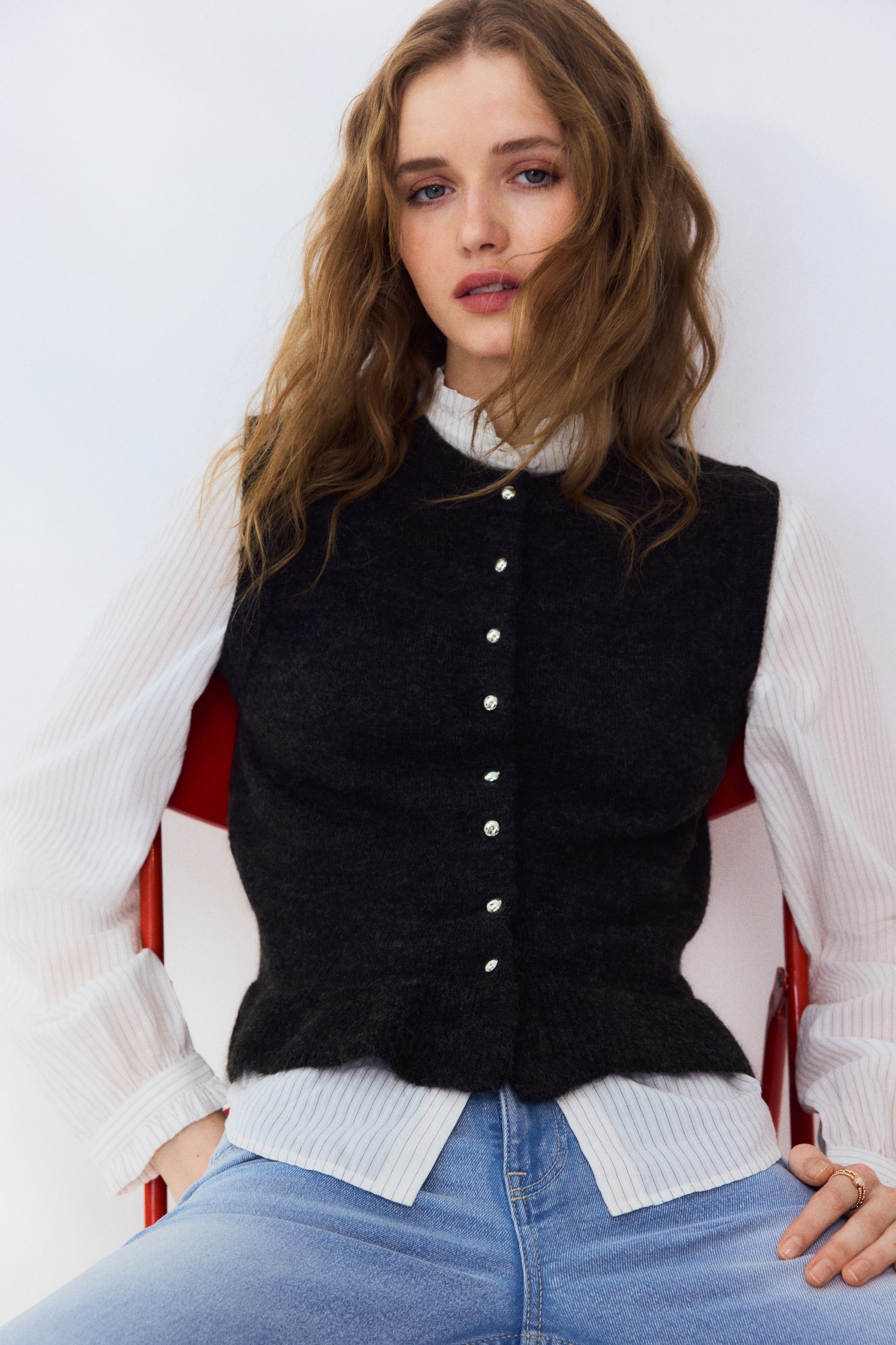 Ampliar la imagen: La modelo lleva puesto un chaleco de punto oscuro y suave sobre una camisa blanca de rayas finas; presenta un escote redondo acanalado, sisas acanaladas, botones brillantes en la parte delantera y un volante acanalado en el dobladillo.