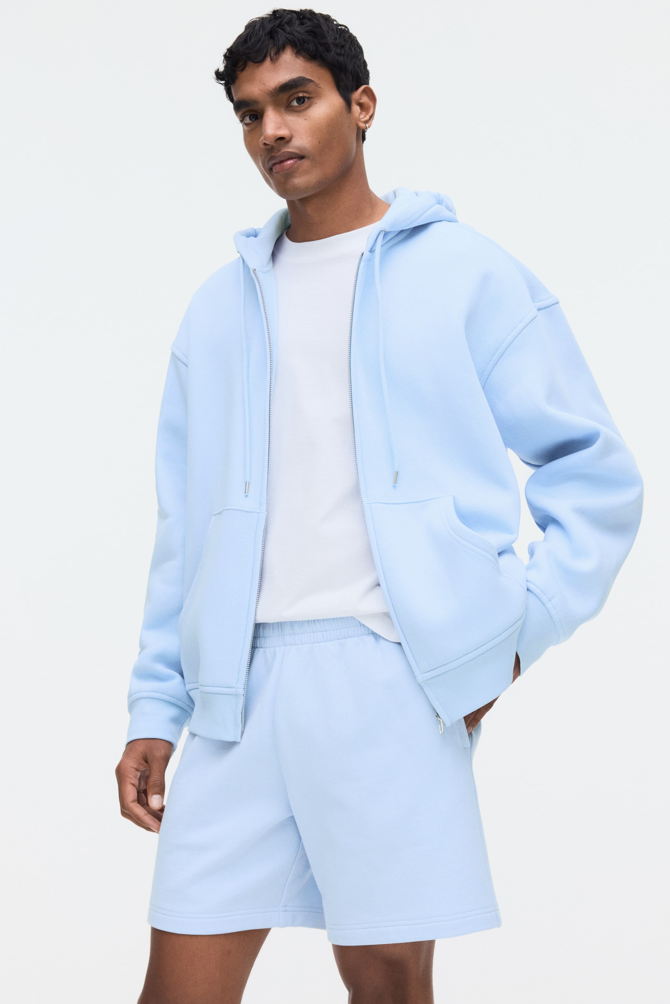 View larger image: The model is wearing light blue shorts made from lightweight cotton sweatshirt fabric. These shorts come with an elasticated waist and side pockets, with the model's hand casually placed in one of the pockets.