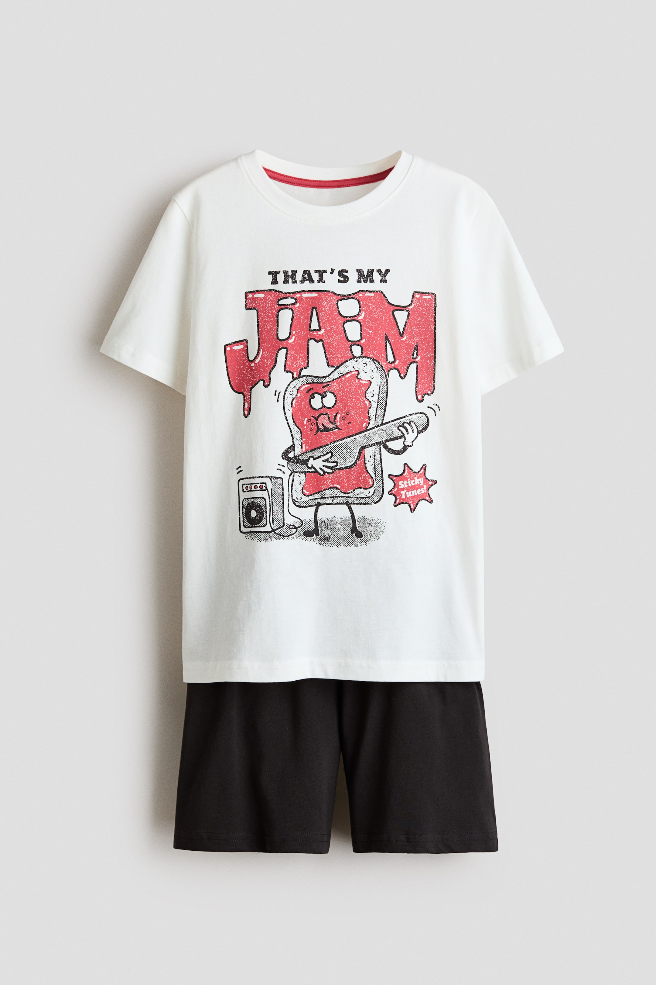 Visualizza immagine più grande: Un set pigiama composto da una t-shirt bianca a maniche corte e pantaloncini neri. La t-shirt presenta un girocollo con bordo interno rosso e una stampa grafica di un personaggio di toast ricoperto di marmellata che suona una chitarra elettrica, con il testo "THAT'S MY JAM" sopra e "Sticky Tunes!" in un'esplosione di stelle.