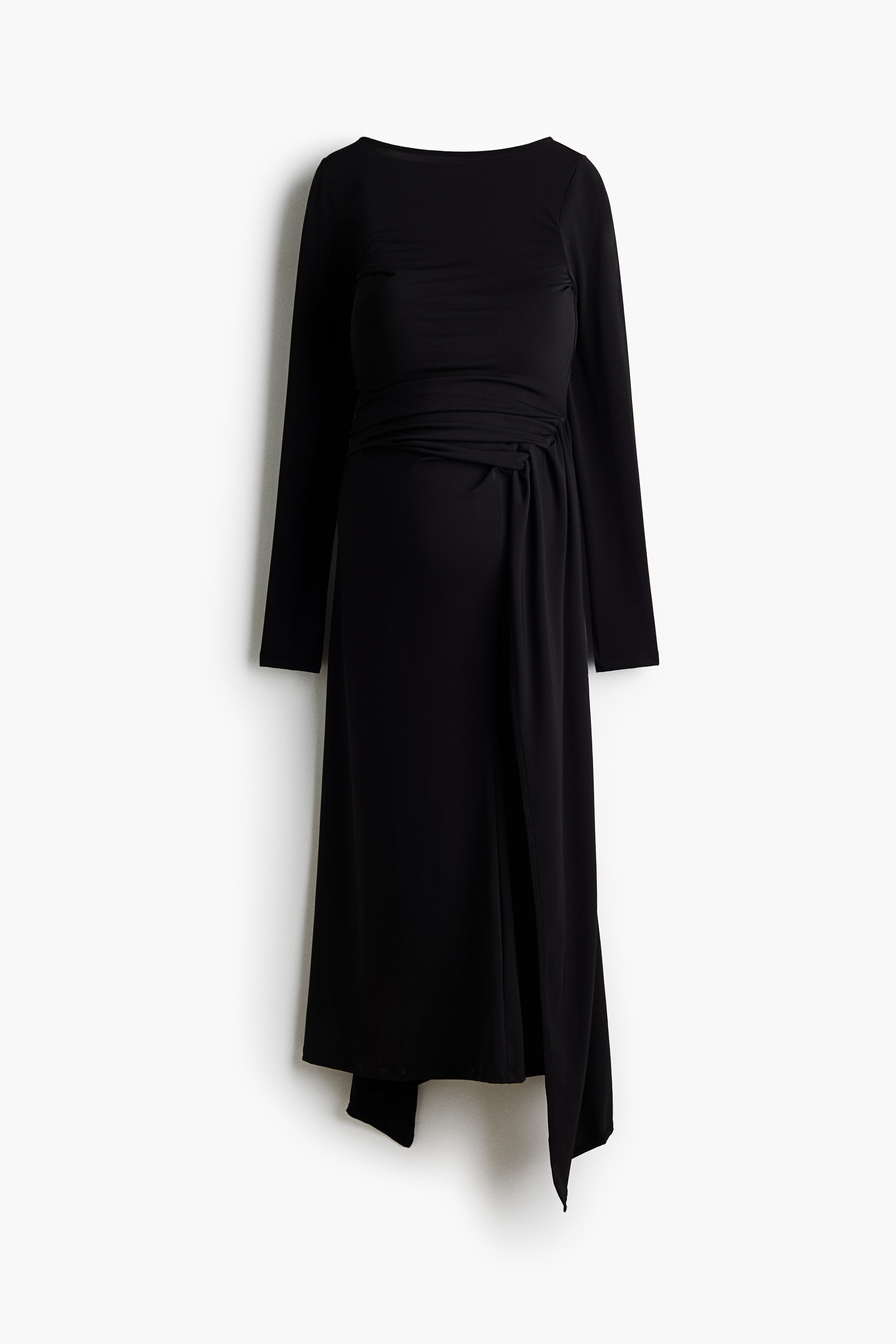 The model is wearing a long, fitted black dress, showcasing its boat neckline, long sleeves and body-skimming silhouette. The dress features wide panels at the hips that meet and tie loosely at the front, creating a draped detail.
