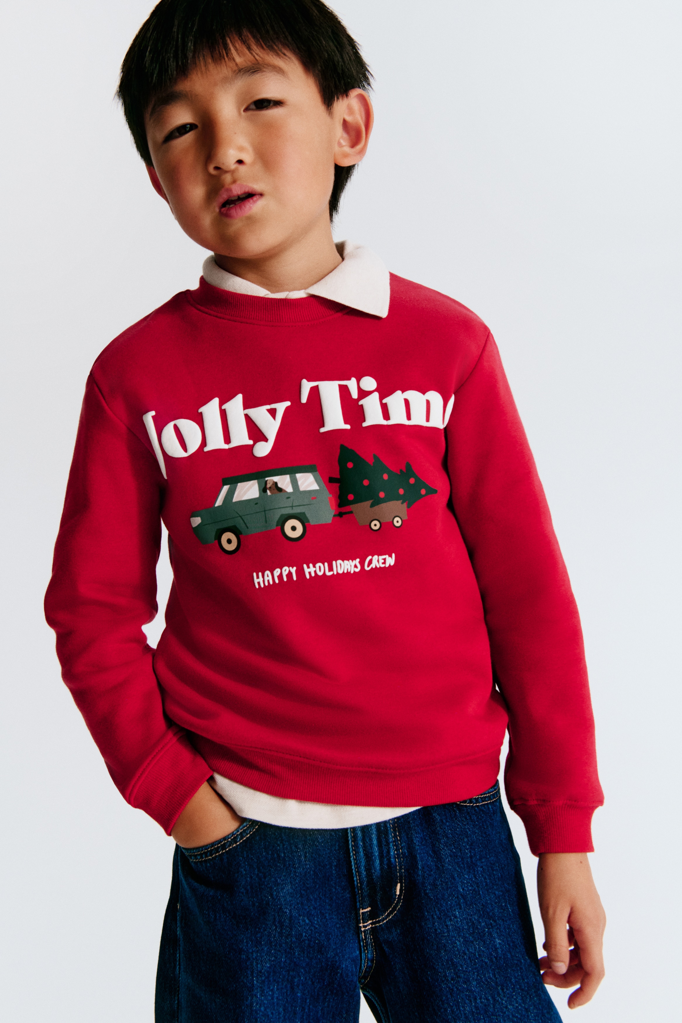 A red sweatshirt is shown from the front, featuring a ribbed round neckline, dropped shoulders, long sleeves, and a ribbed hem and cuffs. The front displays a printed graphic with the white text "Jolly Time" above an illustration of a utility vehicle towing a decorated evergreen tree, with the smaller text "HAPPY HOLIDAYS CREW" printed underneath.