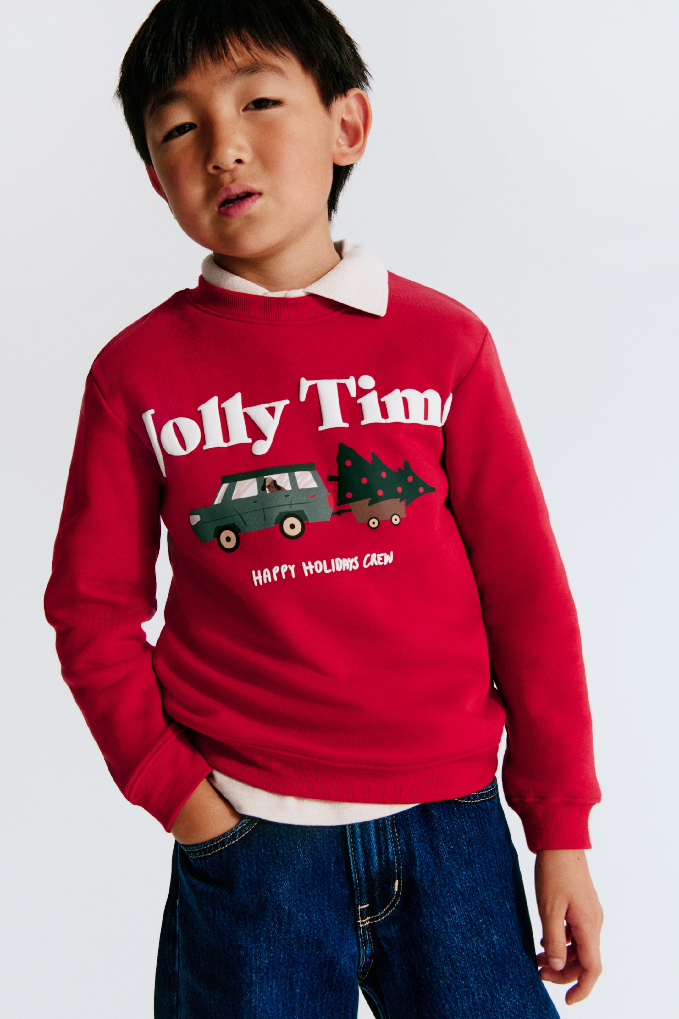 View larger image: A red sweatshirt in sweat fabric is shown from the front, featuring a printed graphic of a green car towing a Christmas tree beneath the white text "Jolly Time." The sweatshirt is designed with a round, rib-trimmed neckline, dropped shoulders, and ribbing at the cuffs and hem, layered over a white collared shirt.