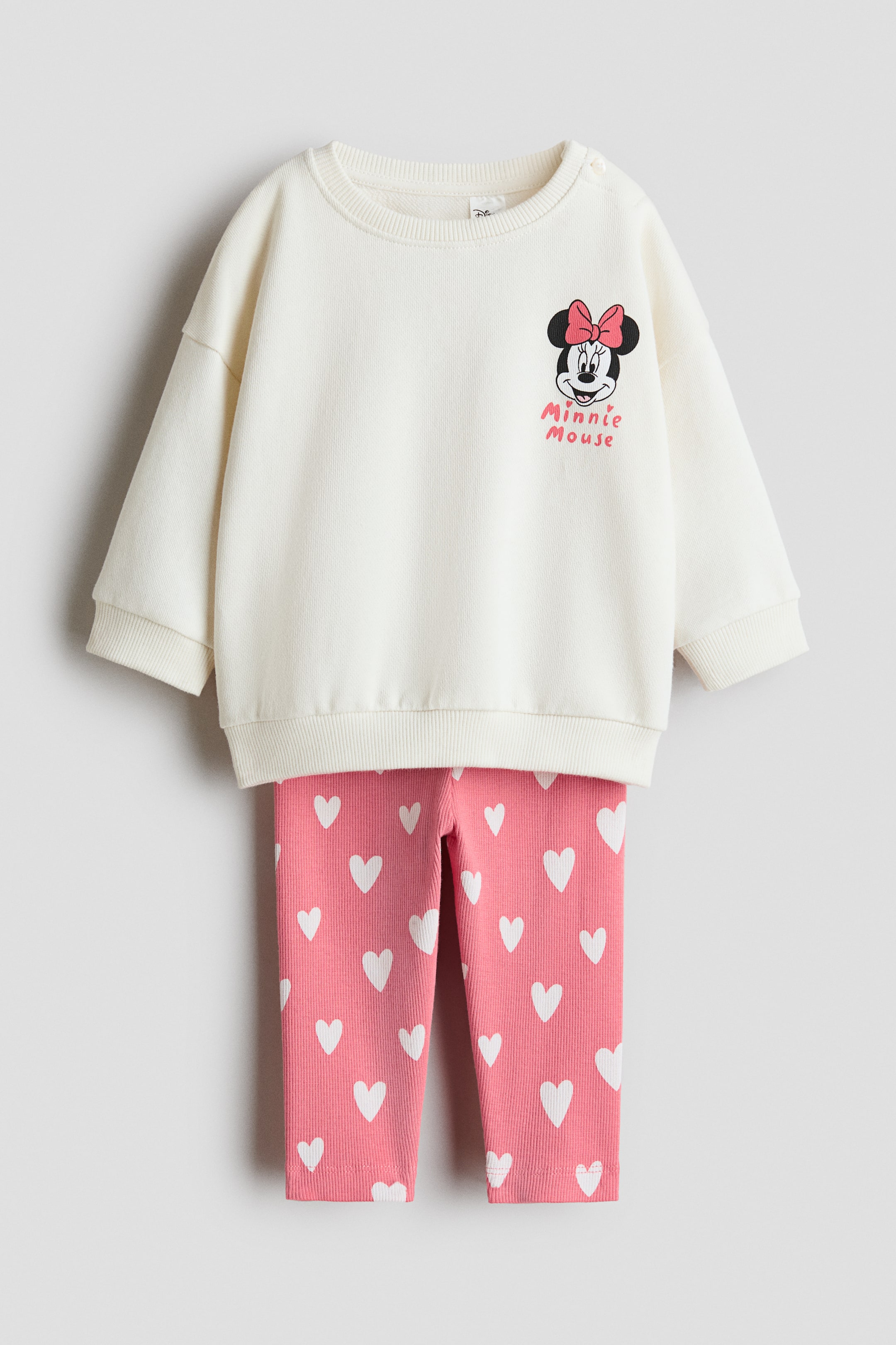 Agrandir l'image: Un ensemble imprimé comprenant un haut à manches longues blanc cassé et un legging rose, présenté de FACE. Le haut affiche un col rond bordé de côtes, un bouton sur une épaule, des poignets côtelés et un ourlet côtelé, avec un graphique de Minnie Mouse et du texte sur la poitrine. Le legging rose présente un imprimé de cœurs blancs sur l'ensemble et une taille élastiquée.