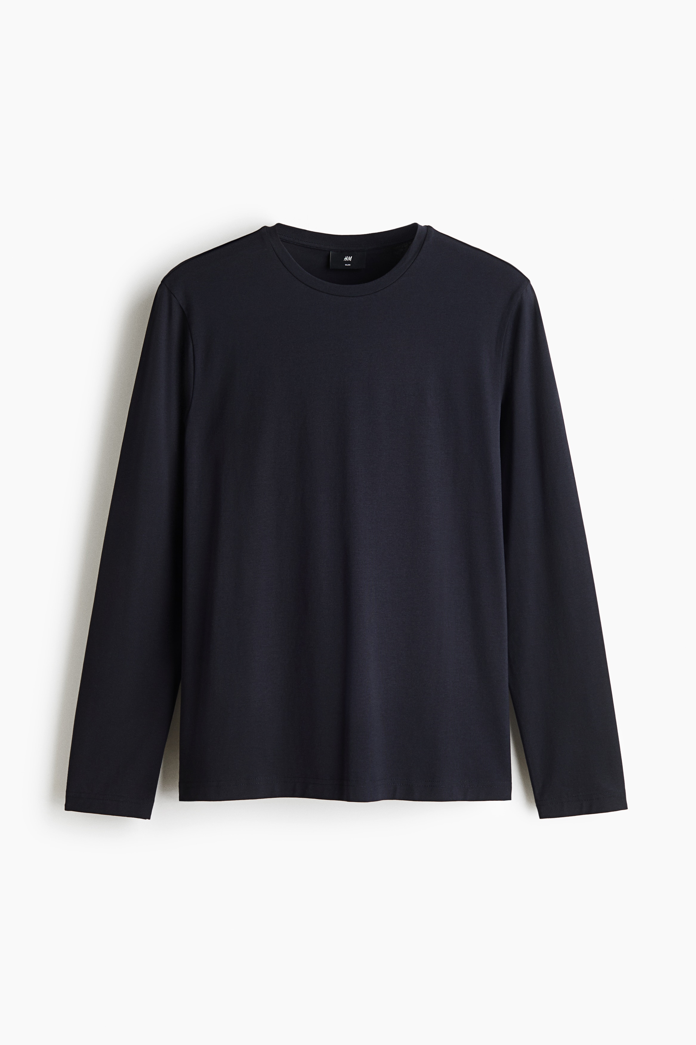 Das Model trÃ¤gt ein langÃ¤rmeliges T-Shirt mit Rundhalsausschnitt in dunklem Marineblau, kombiniert mit einer locker geschnittenen dunklen Hose. Eine cremefarbene Canvas-Tote-Bag wird in einer Hand getragen und rundet das Outfit ab.