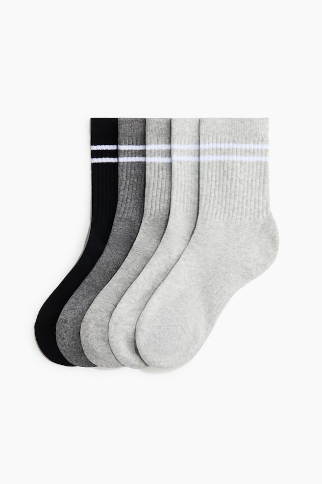 Cinco calcetines deportivos tipo crew aparecen superpuestos en una vista de perfil, mostrando una gradaciÃ³n de colores del negro al gris claro. Cada calcetÃ­n presenta una secciÃ³n de caÃ±a acanalada con dos franjas horizontales blancas cerca del borde elÃ¡stico y un pie de punto fino. - 1