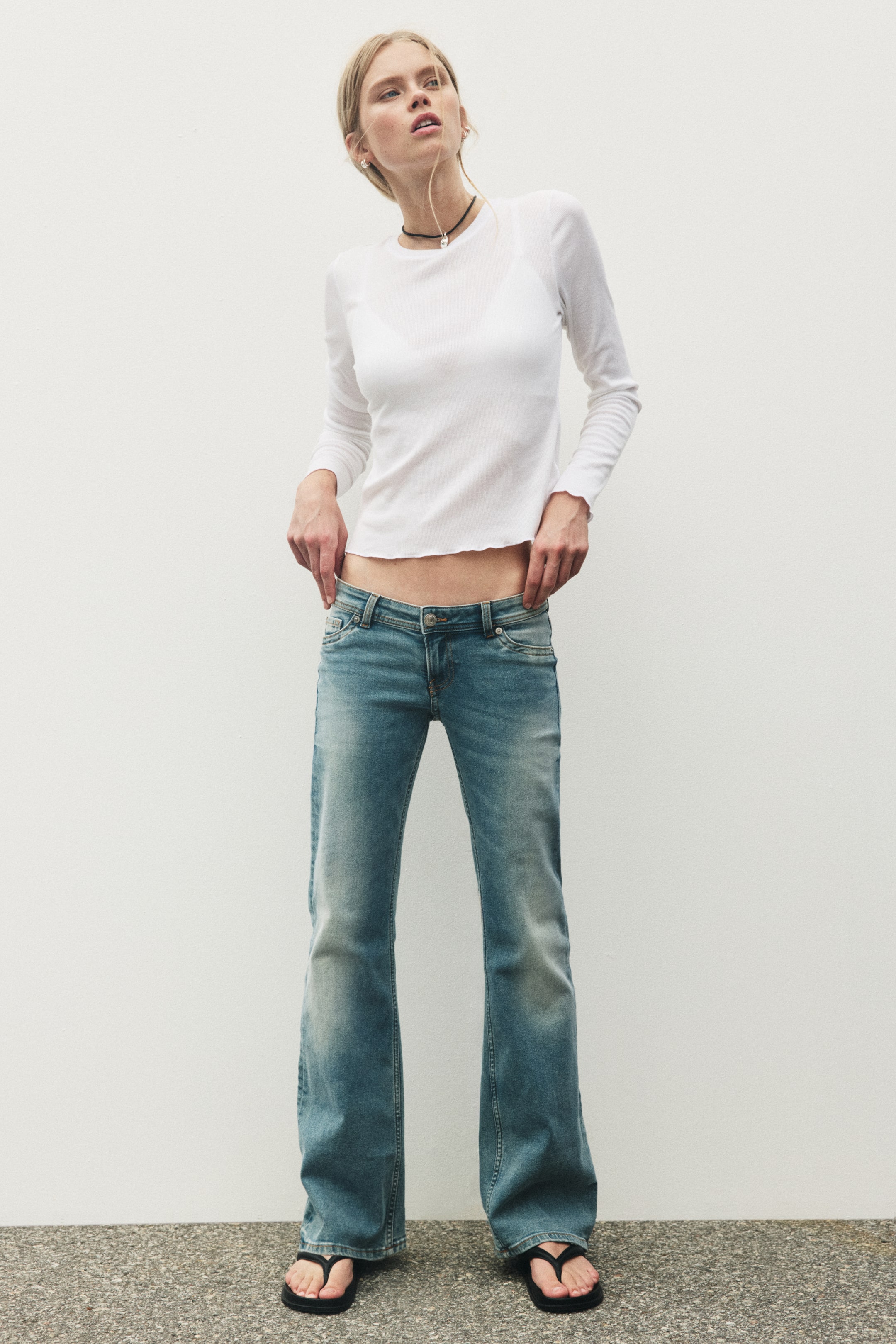 Visa större bild: Jeans i denim med extra låg midja och utsvängda ben bärs av modellen. Jeansen har knappstängning, myntficka och öppna bakfickor, med fållen som slutar vid fotens ovansida, medan modellens händer håller i midjan.