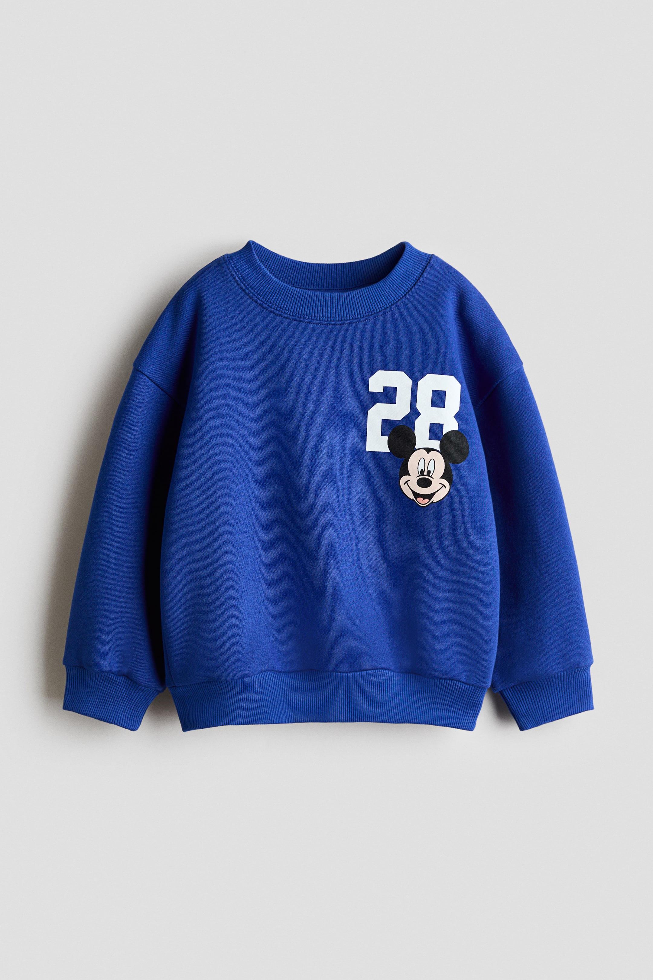 Se større billede: En blå sweatshirt vises forfra med rund ribkantet halsudskæring, sænkede skuldre samt ribkanter ved ærmer og nederst. Forstykket har et hvidt trykt motiv af tallet "28" med en Mickey Mouse-illustration, der overlapper nederste del.