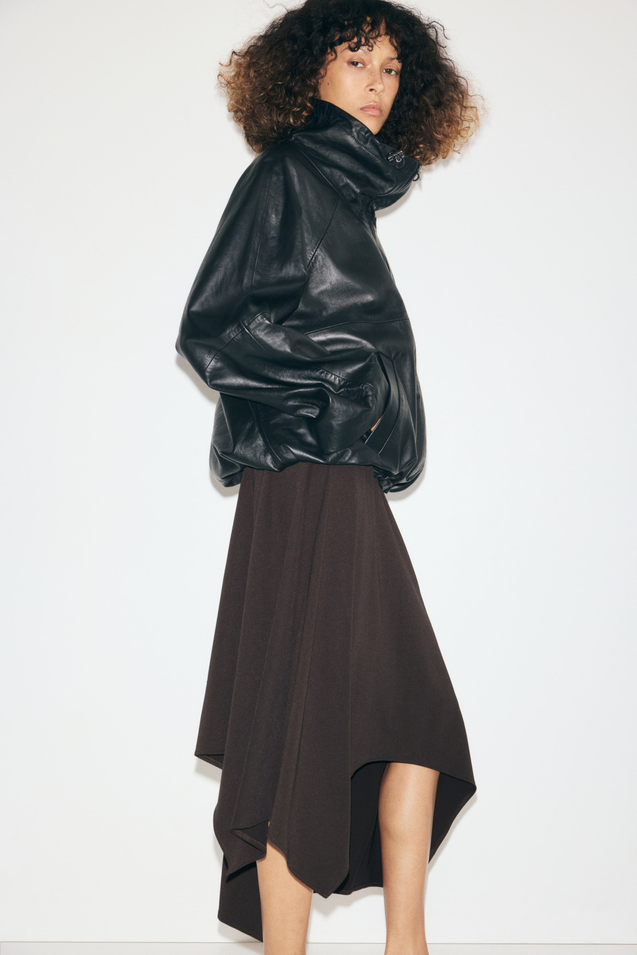 View larger image: A dark brown, calf-length skirt in textured stretch jersey is worn by the model, featuring a loose A-line fit and an asymmetrical handkerchief hem. The skirt is styled with a high-collared black leather jacket layered over the waist.