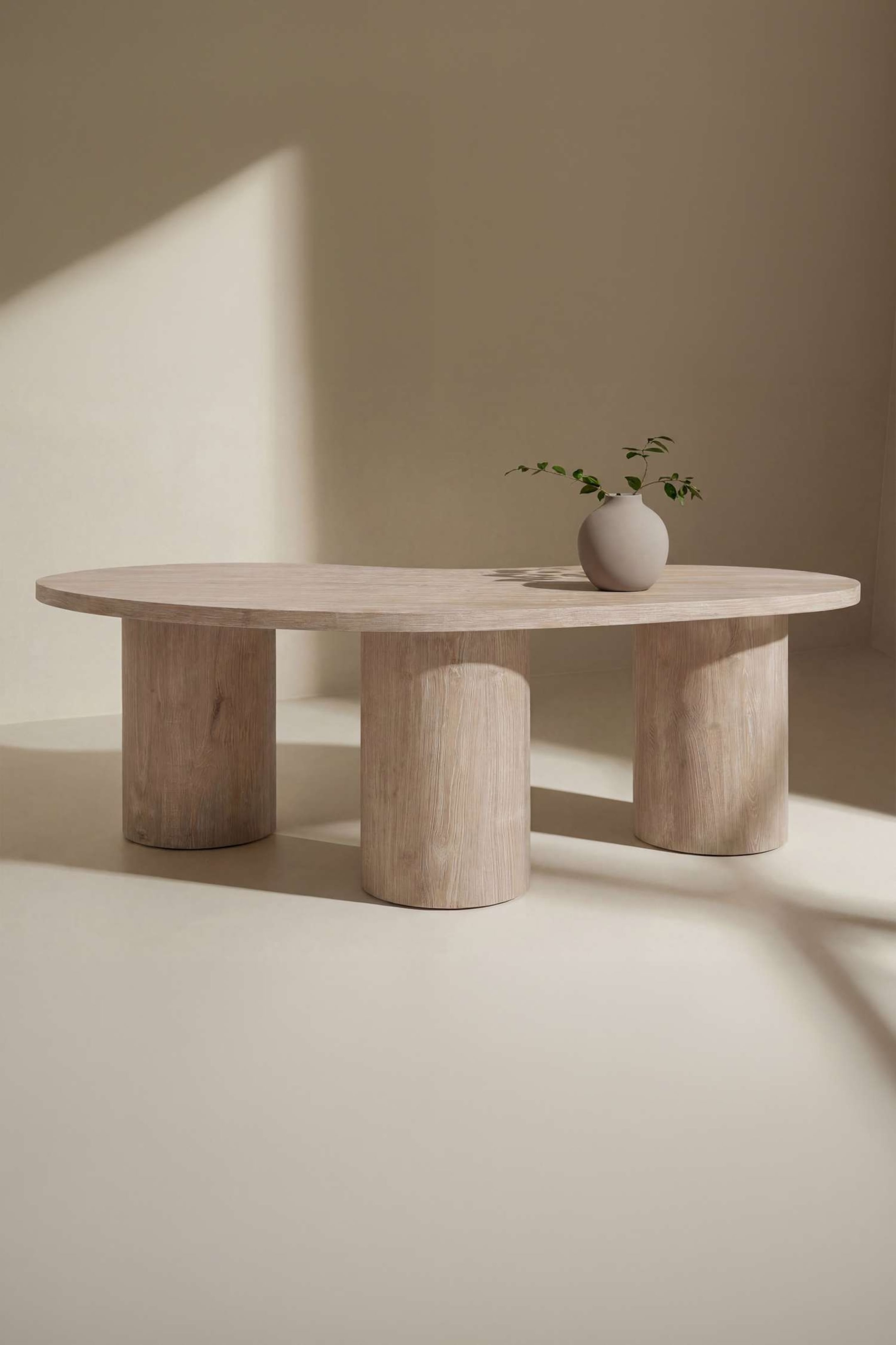 Grotere afbeelding bekijken: De ovale eettafel heeft een lichte houtafwerking met zichtbare nerfstructuur en wordt ondersteund door drie cilindervormige kolompoten. Op het blad staat een kleine grijze vaas met groen blad.