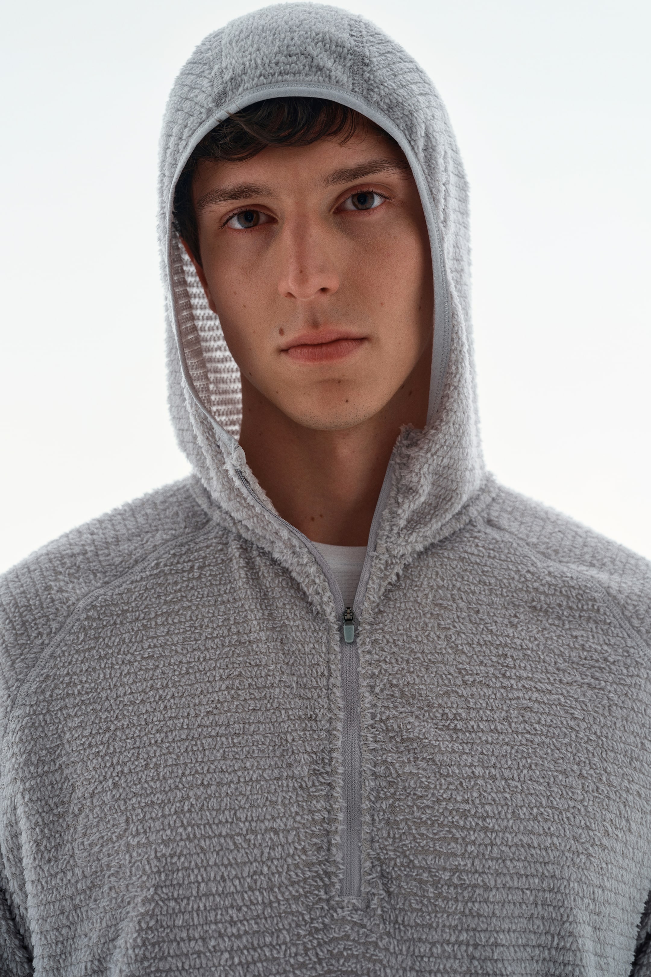 Xem ảnh lớn hơn: Một chiếc áo hoodie vải nỉ có kết cấu, màu xám nhạt, với thiết kế khóa kéo nửa thân đang được người mẫu mặc, mũ trùm đầu kéo lên. Khóa kéo được mở một phần, để lộ phần cổ áo thun trắng bên trong.