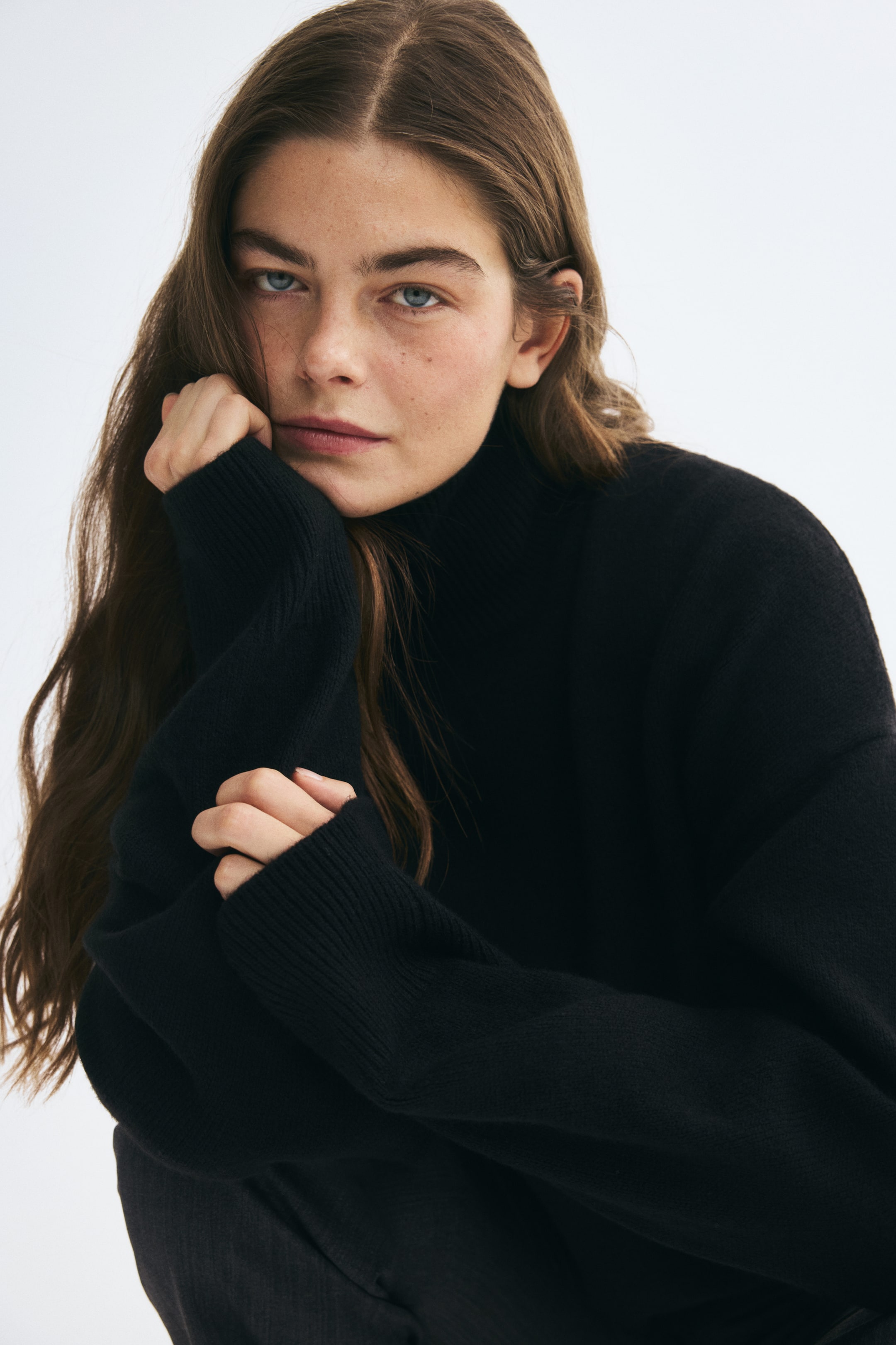 Agrandir l'image: Un pull noir oversize en maille douce, doté d’un col montant côtelé et de longues manches avec poignets côtelés, est porté par le mannequin. Le mannequin pose son menton sur une main, mettant en valeur les détails côtelés de la manche.