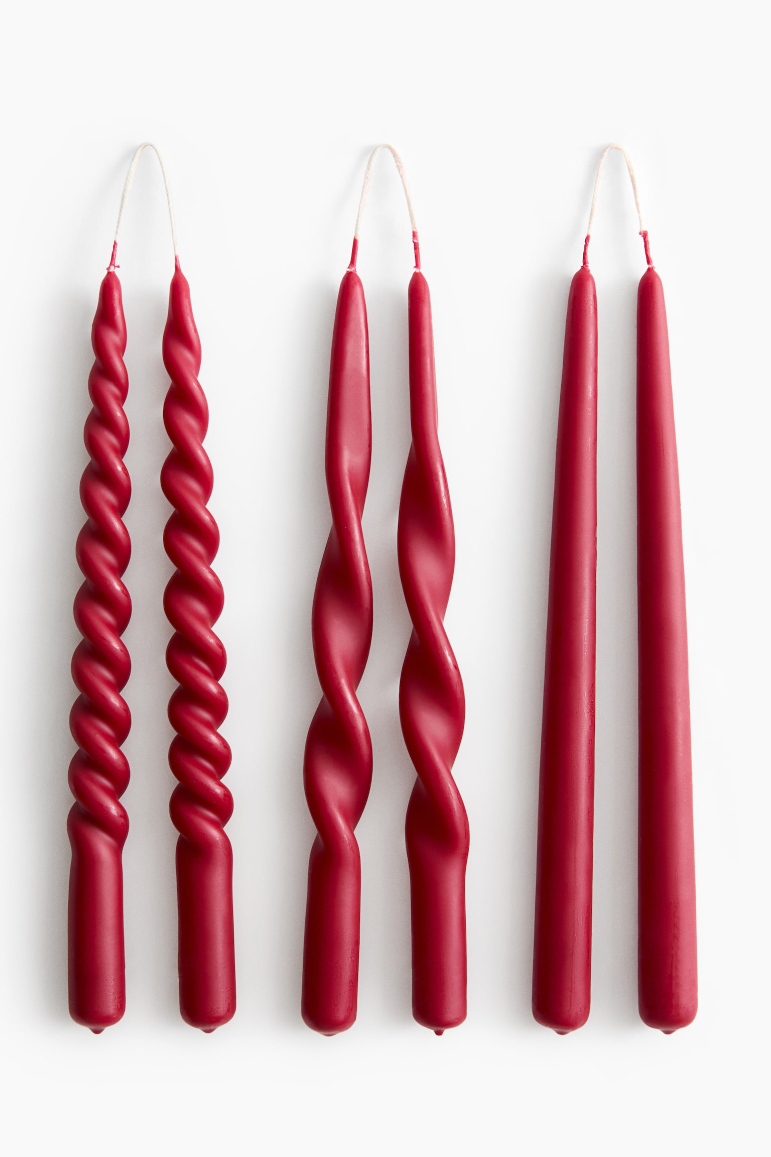 Tres pares de velas cónicas rojas cuelgan una al lado de la otra de mechas continuas, mostrando tres formas distintas de izquierda a derecha: una espiral ajustada, una espiral suelta y una clásica vela cónica lisa.