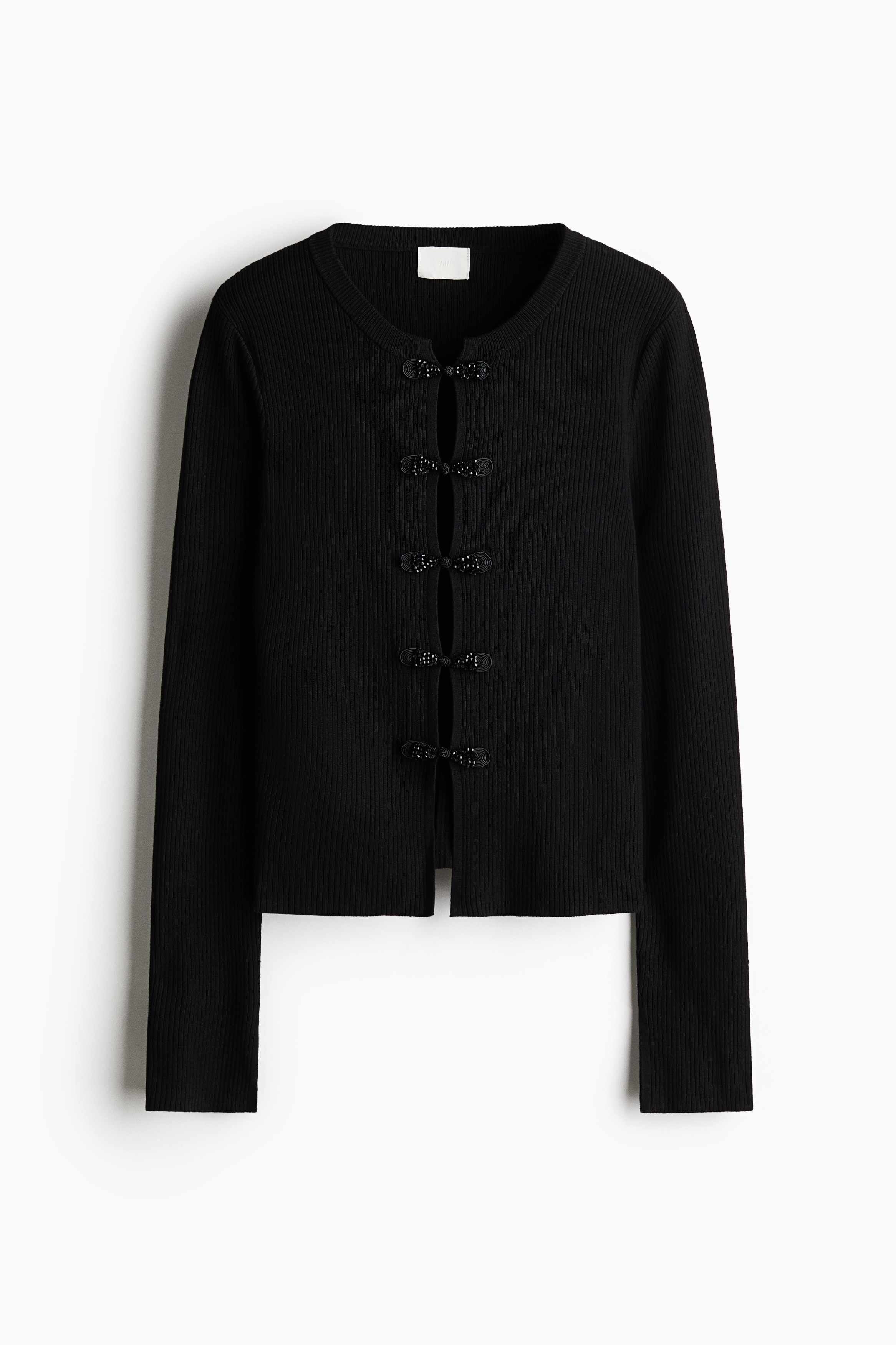 The model is wearing a black, fitted rib-knit cardigan paired with a matching black rib-knit skirt. The cardigan features long sleeves, a round neckline and a front fastening secured with black knot buttons with beaded ends, shown partially unfastened as the model poses with one hand on their hip and the other resting near their neck.