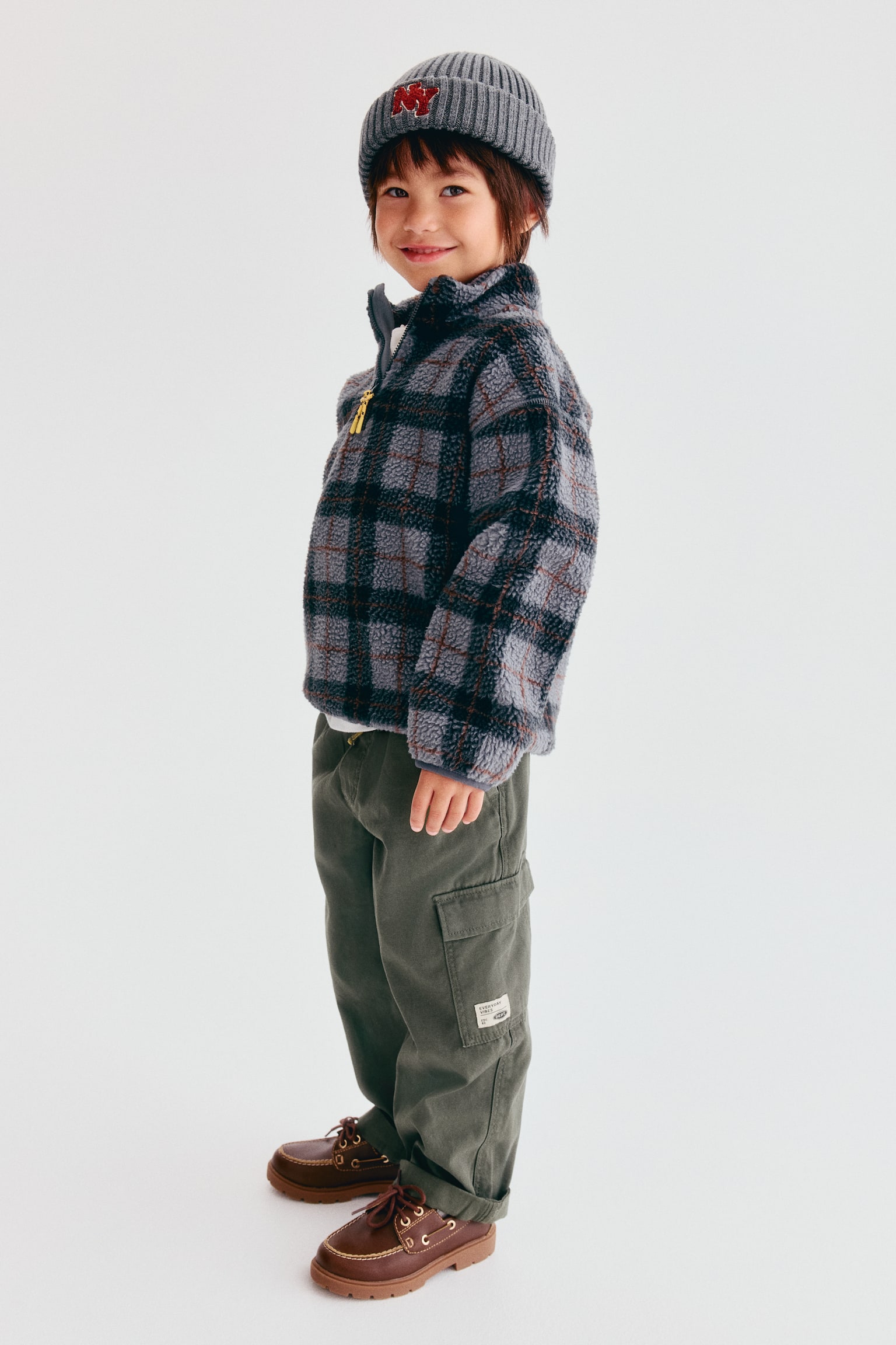 Cargo joggers in dark green cotton canvas are shown from a side-facing angle, highlighting their loose fit and utility design. The joggers feature a diagonal side pocket and a patch leg pocket with a flap on the side of the leg. - 1