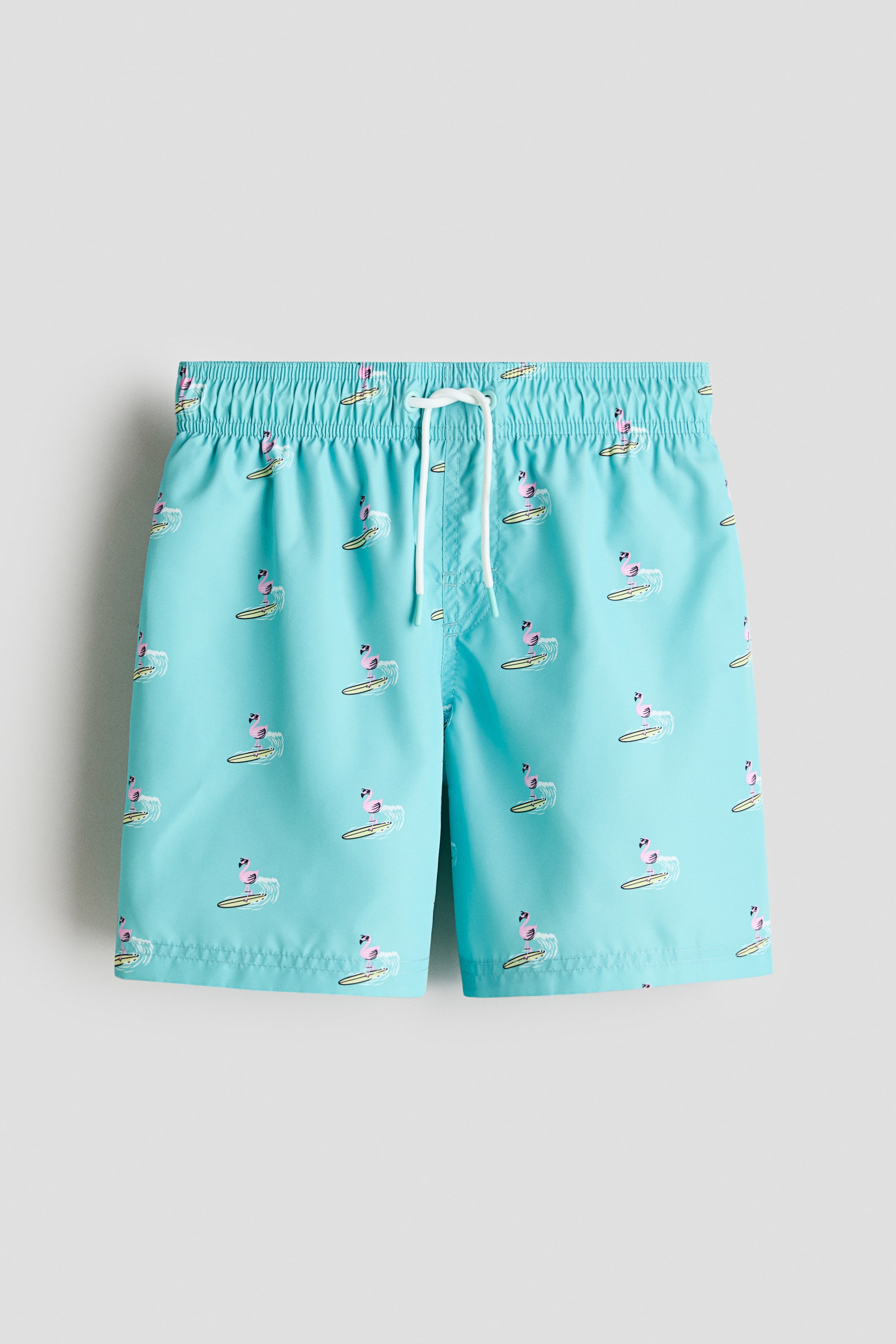 View larger image: The aqua blue swim shorts boast an all-over print of pink flamingos surfing, complete with an elasticated waist and a white drawstring tied at the front.
