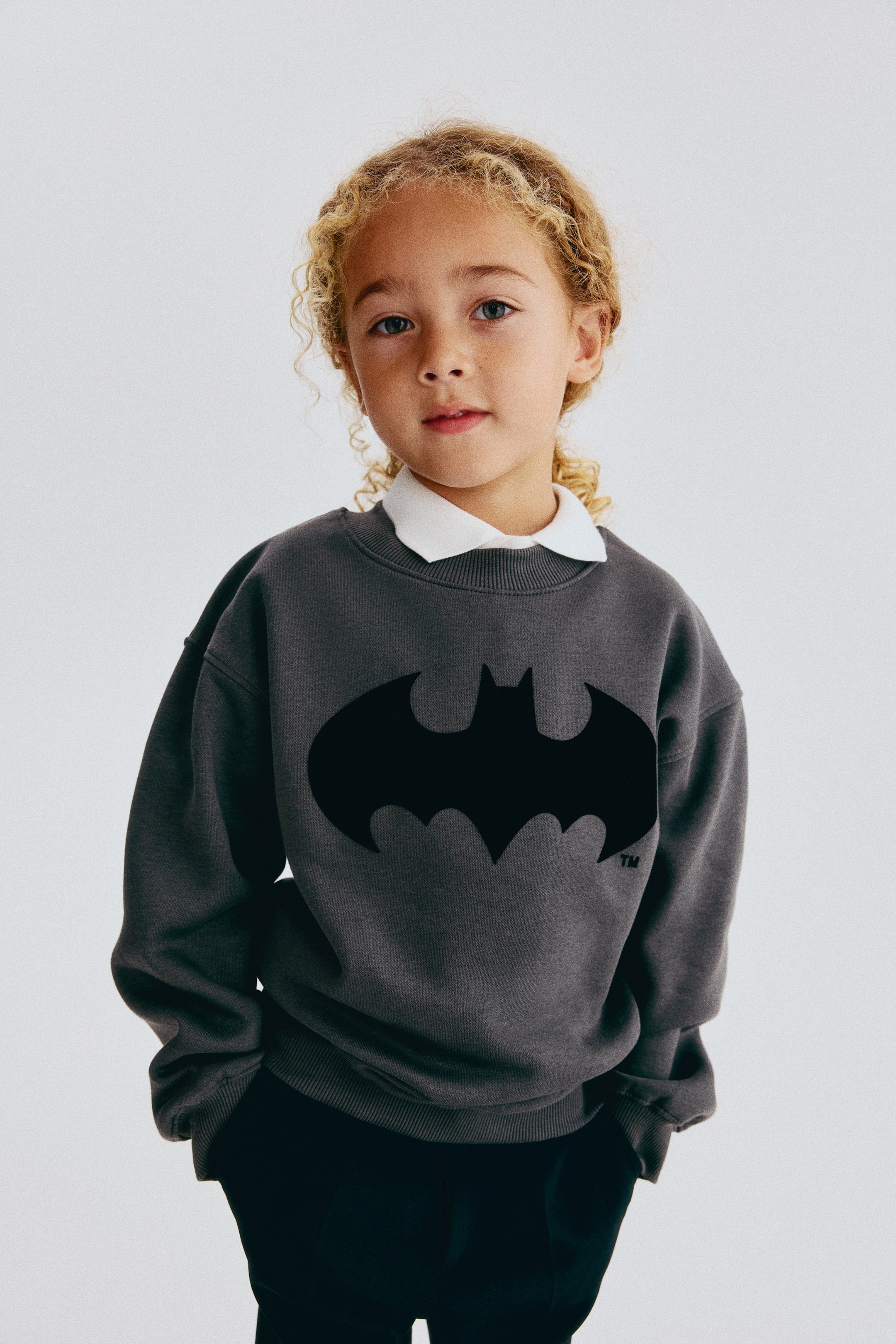View larger image: A dark grey sweatshirt featuring a black Batman logo on the chest is shown from a front angle, showcasing its relaxed fit with dropped shoulders, a rib-trimmed crew neck, and a ribbed hem. The sweatshirt is styled layered over a white collared shirt.