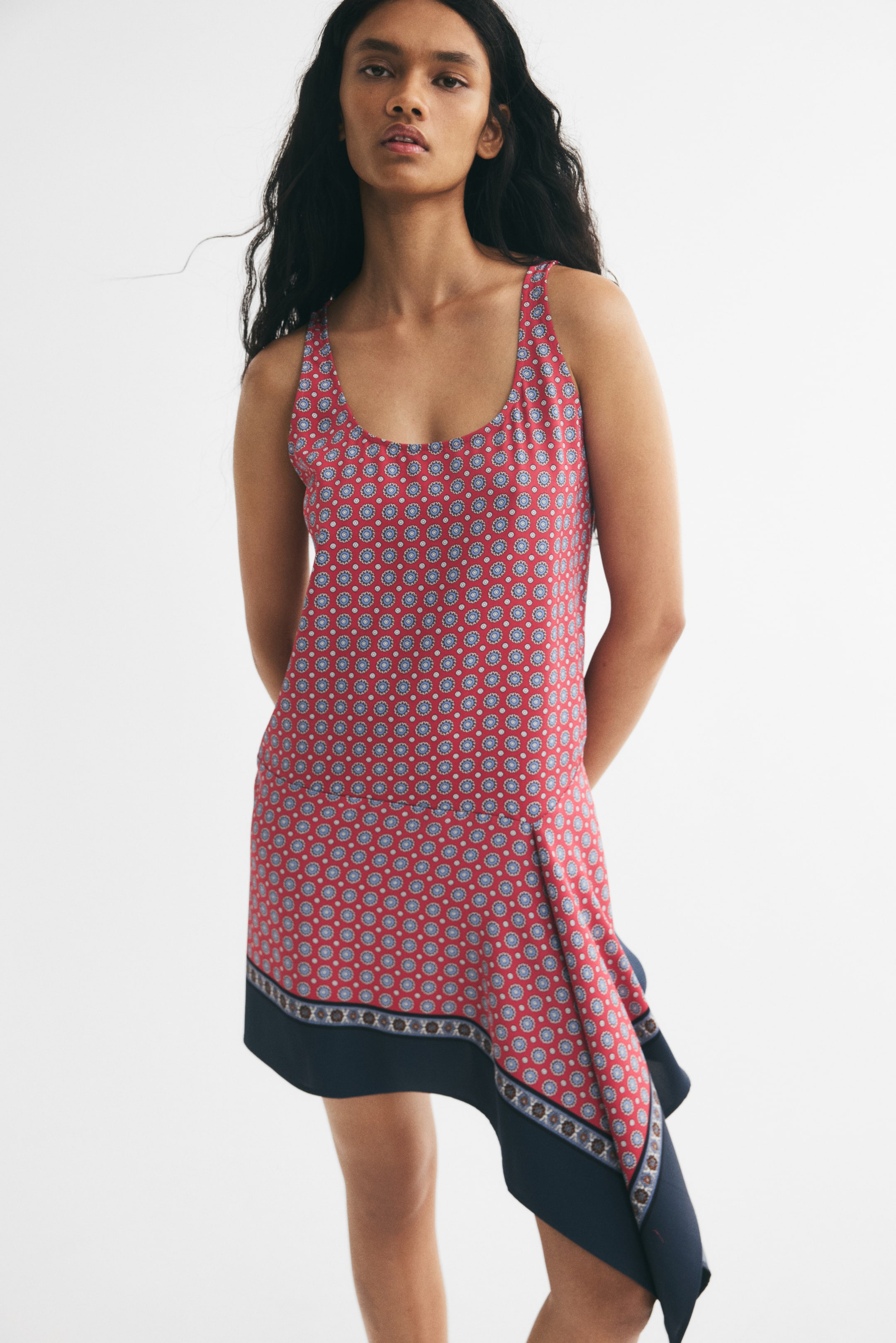 Ampliar la imagen: La modelo lleva puesto un vestido corto sin mangas de sarga con un patrón geométrico en rojo y azul. El vestido está diseñado con un escote redondo, una cintura de corte bajo y un dobladillo asimétrico con un panel drapeado azul oscuro en la parte frontal.