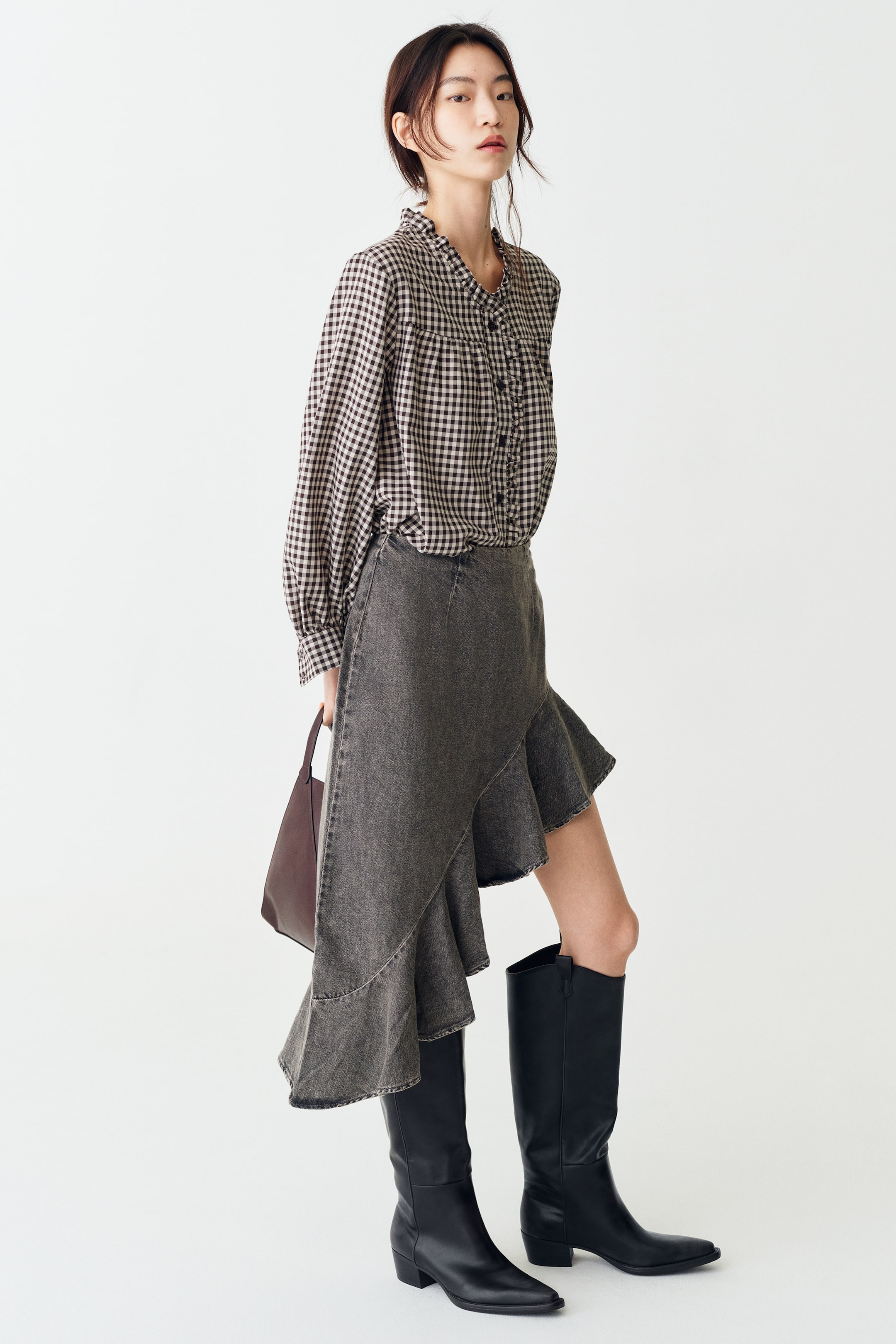 Xem ảnh lớn hơn: Người mẫu đang mặc một chiếc áo blouse vải twill họa tiết gingham, nổi bật với cổ bèo nhún và đường viền bèo nhún đồng bộ dọc theo thân trước cài nút. Áo có tay dài phồng, bo cổ tay nhún và được sơ vin vào chân váy denim xám bất đối xứng.
