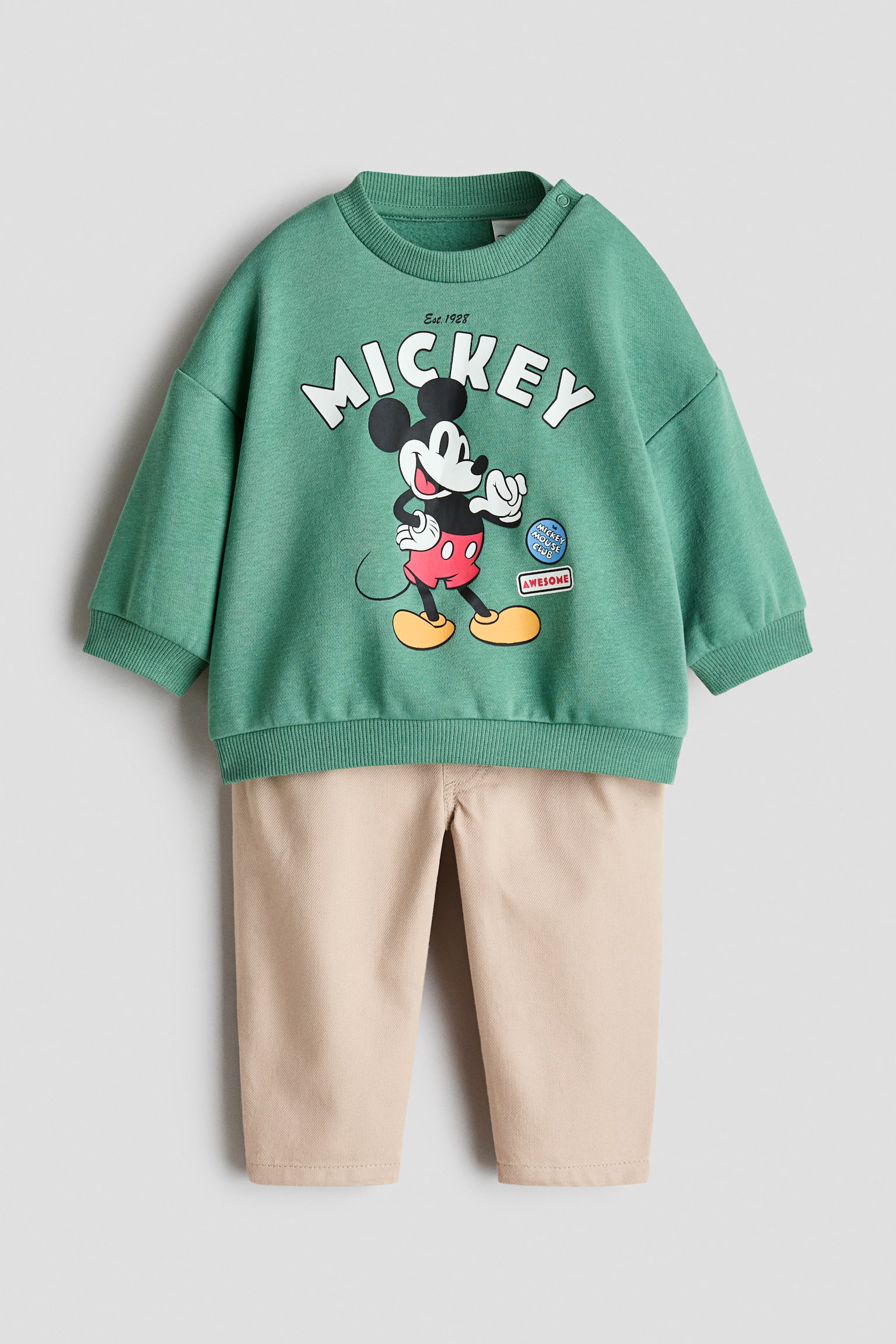 A two-piece childrenâs outfit, shown from the front, features a green sweatshirt and beige trousers. The green sweatshirt displays a central Mickey Mouse graphic beneath the word âMICKEYâ, along with dropped shoulders, a ribbed crew neck with a snap fastener, ribbed cuffs and a ribbed hem, while the coordinating beige trousers have a straight-leg fit.