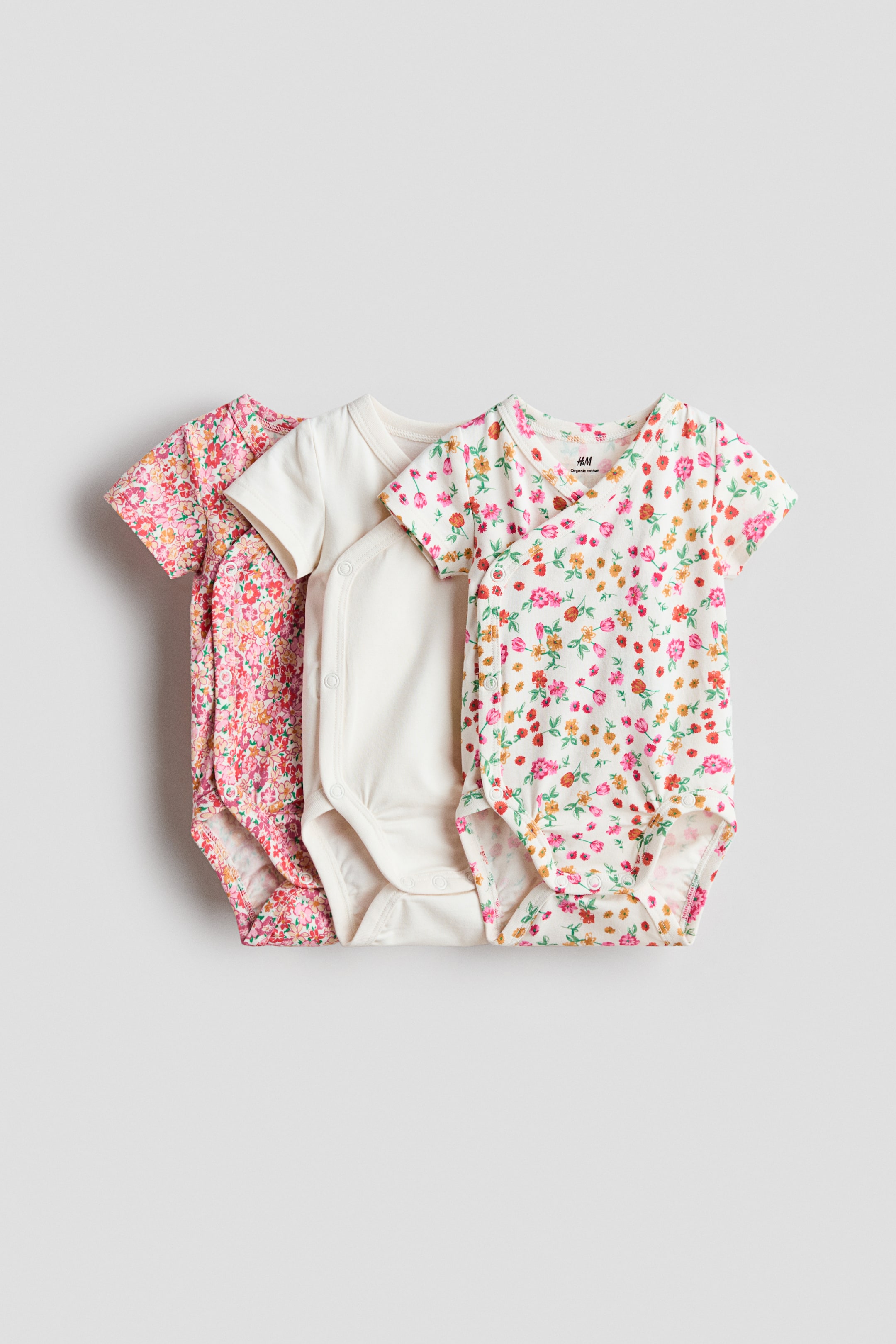 View larger image: Three short-sleeved, wrapover bodysuits crafted from soft cotton jersey are showcased, highlighting their front designs. These bodysuits feature visible press-studs along one side and at the crotch for convenience. The collection includes a pink floral print, a solid cream, and a white bodysuit with a multi-color floral print.