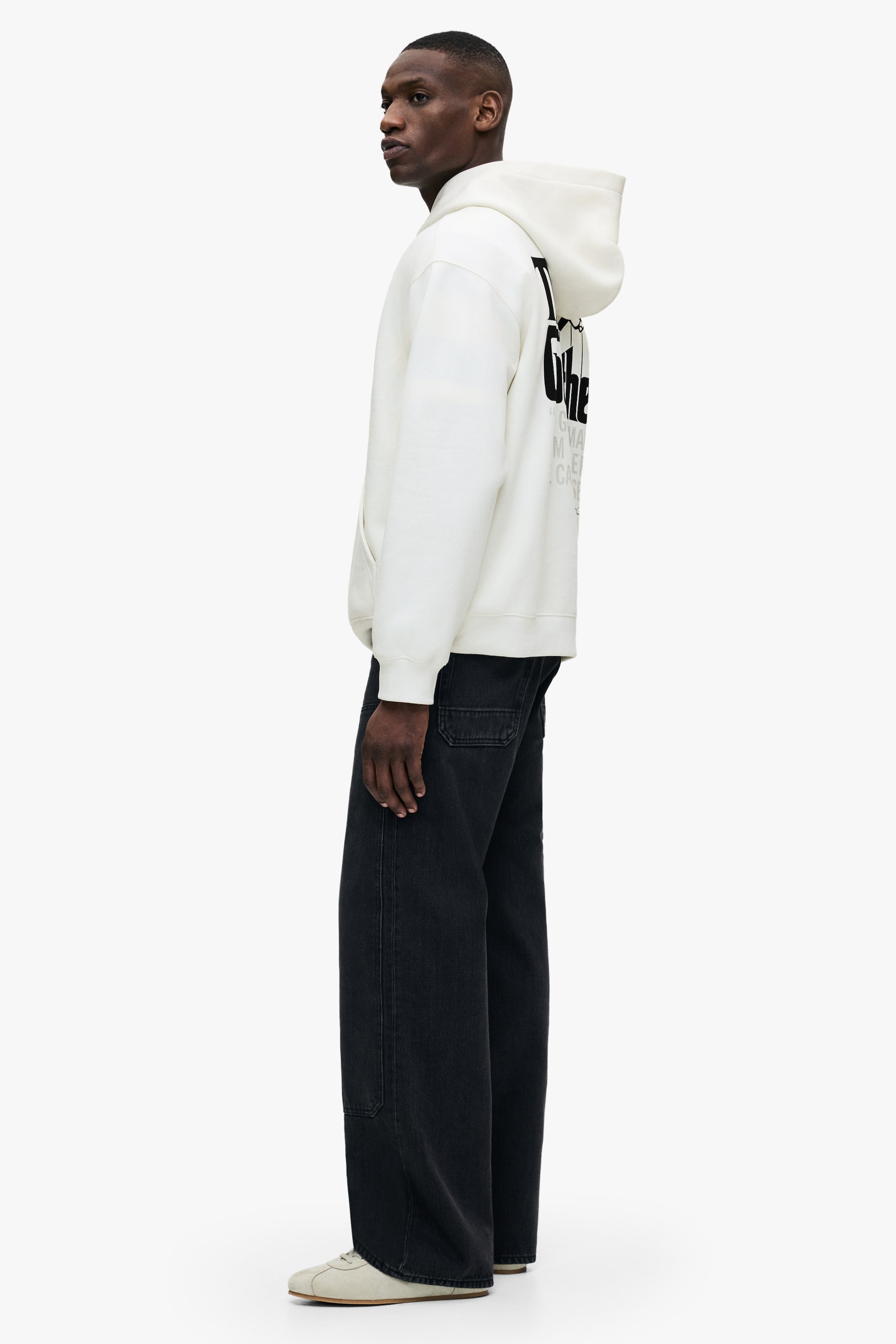 拡大画像を表示: 白いフーディーがモデルによって着用されており、背中には黒のグラフィックプリントが施されています。それにはフードとフロントのカンガルーポケットがあり、暗めのジーンズと明るい色のスニーカーとスタイリングされています。