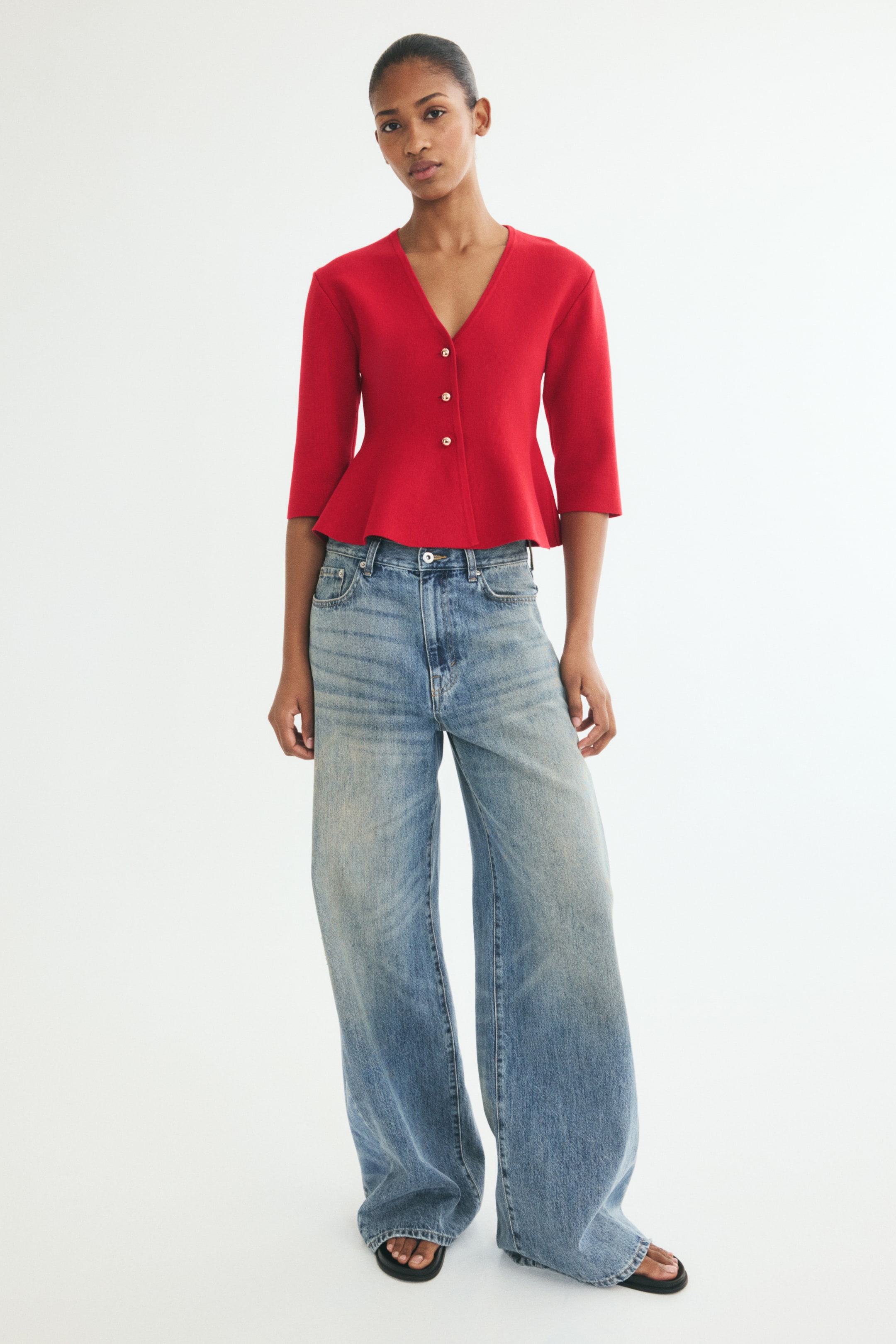 View larger image: A red, high-density knit cardigan is shown from the front, featuring a fitted silhouette with a deep V-neckline and three small metallic buttons down the front. The cardigan displays slightly dropped shoulders, three-quarter-length sleeves, and a flared peplum hem.