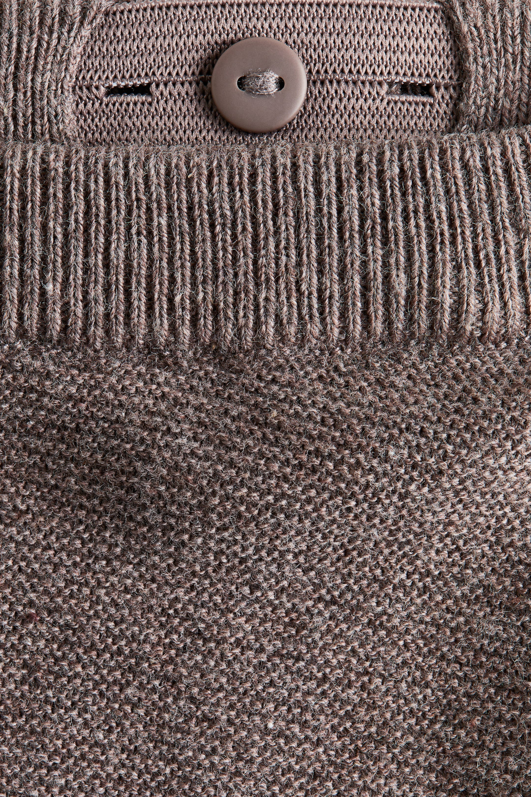 Grösseres Bild ansehen: Eine Nahaufnahme zeigt einen braunen H&M Adorables Pullover, der aus einem strukturierten Patentstrickgewebe besteht, das in ein geripptes Strickband übergeht. Ein brauner Knopf ist auf einer oberen Strickleiste sichtbar, neben einem offenen Knopfloch positioniert, was auf die Rückenhalsöffnung des Pullovers hinweist.