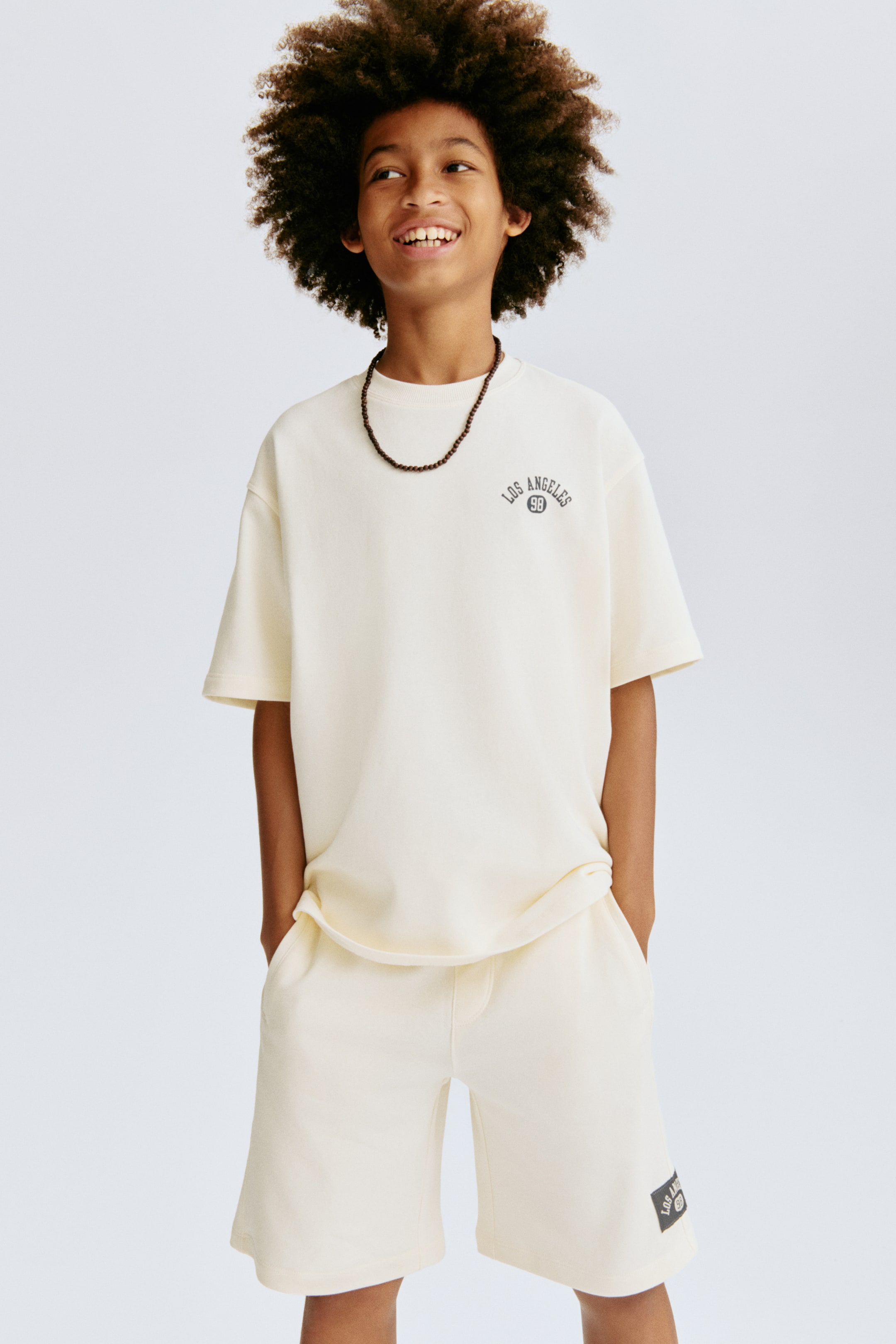 Ampliar la imagen: Un conjunto de camiseta de manga corta de ajuste relajado y color crema con shorts a la altura de la rodilla se muestra desde un ángulo frontal. La camiseta presenta un cuello redondo y el texto "LOS ANGELES 98" en el pecho izquierdo, mientras que los shorts tienen un parche de logotipo coordinado en la pierna izquierda.