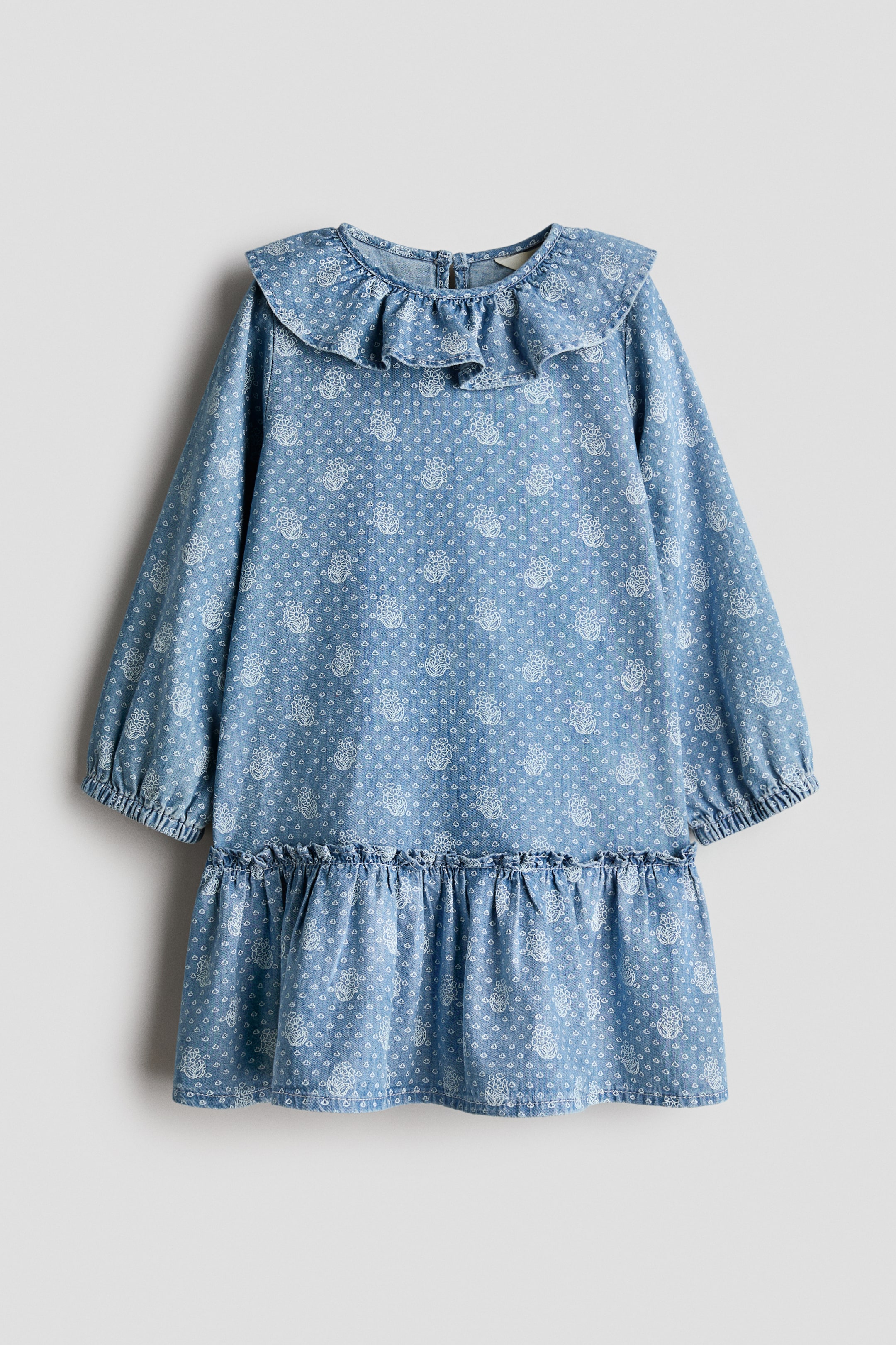 Agrandir l'image: Une robe en chambray bleu clair vue de face met en valeur un imprimé floral délicat blanc sur l’ensemble, avec un large col à volants, des manches longues à poignets élastiqués et un ourlet froncé à volants.